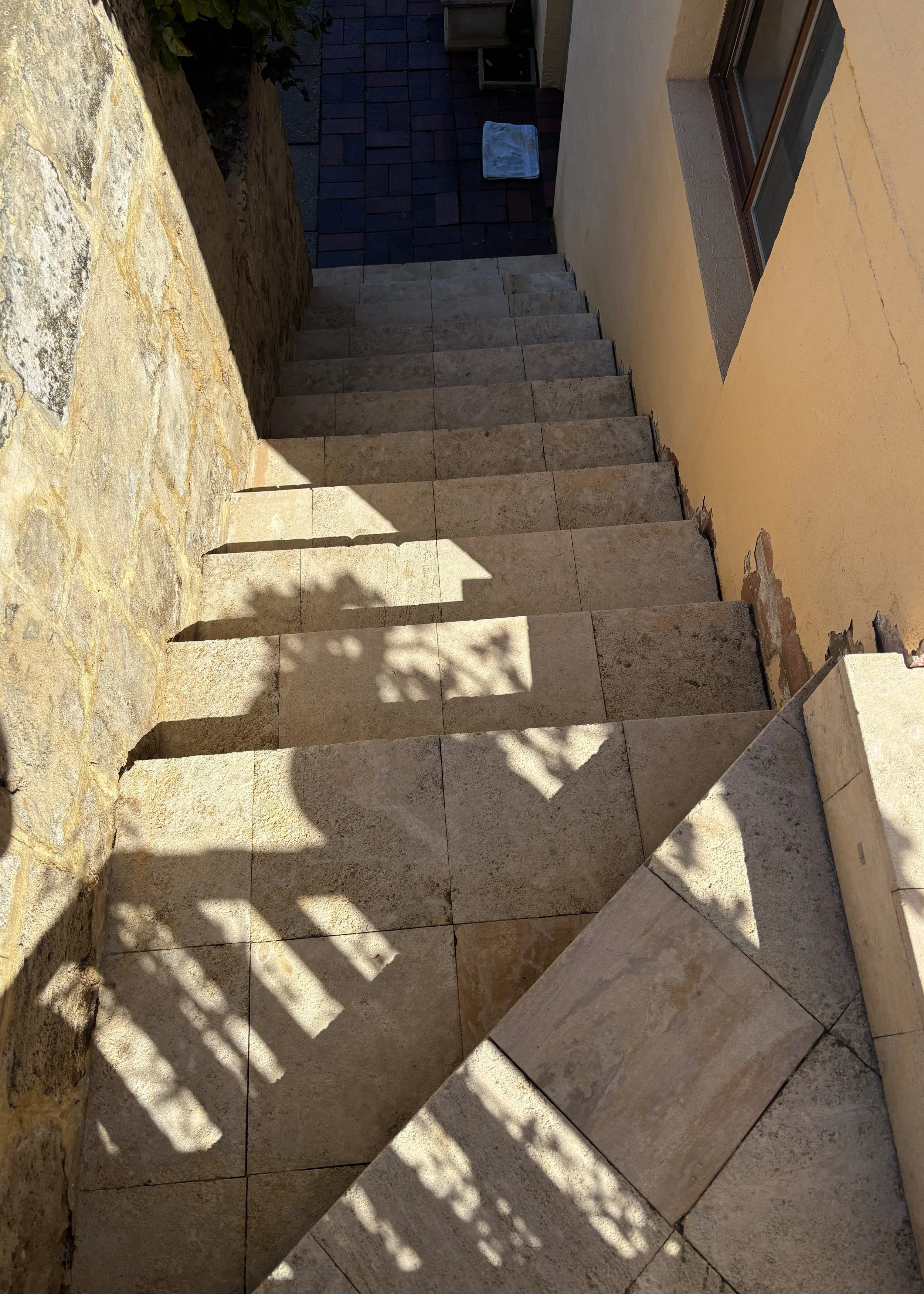 An outdoor staircase leading down from a house with yellow walls, with shadows of a fence and a plant cast on the stairs, and a window on the right side.