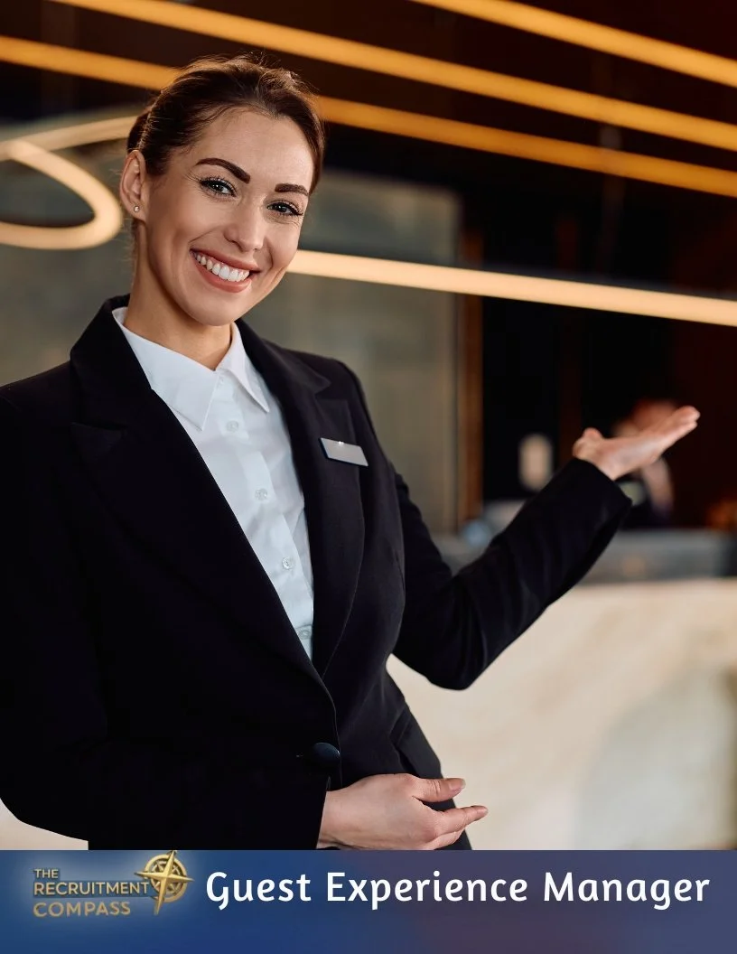 A smiling woman in a black suit and white shirt at a reception desk, gesturing with her right hand, with a professional background. The text on the image reads "The Recruitment Compass Guest Experience Manager."