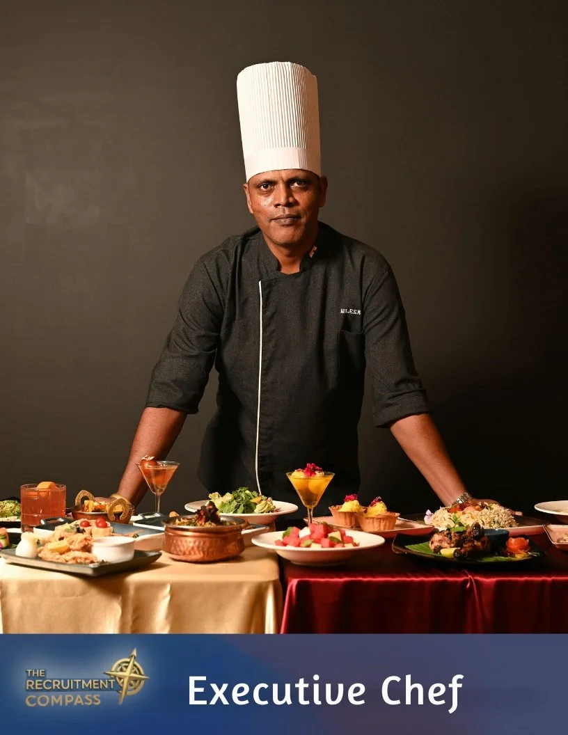 A chef in a black uniform and tall white chef hat stands behind a table with various plated dishes and desserts, against a dark background. The text overlay reads 'Executive Chef' and 'The Recruitment Compass'.