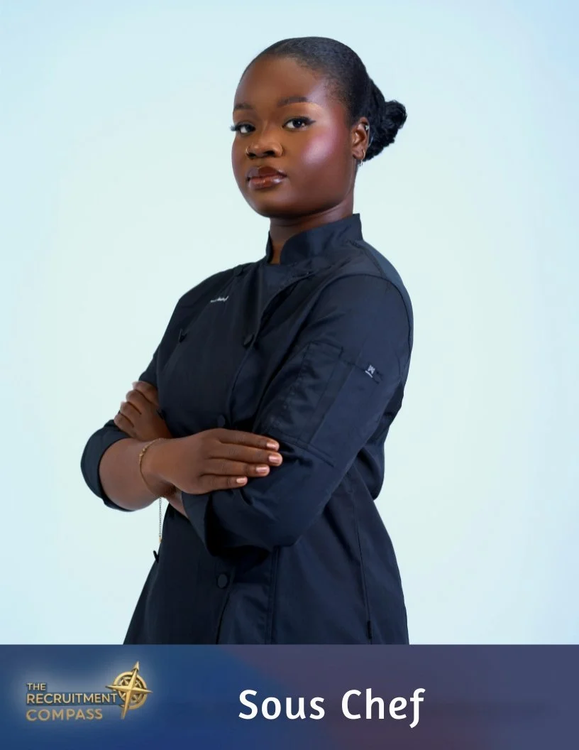 A young woman with dark skin and curly hair tied back, wearing a black chef's coat, standing with arms crossed against a light background. The lower part of the image has a banner with the text 'Sous Chef' and a logo for 'The Recruitment Compass'.