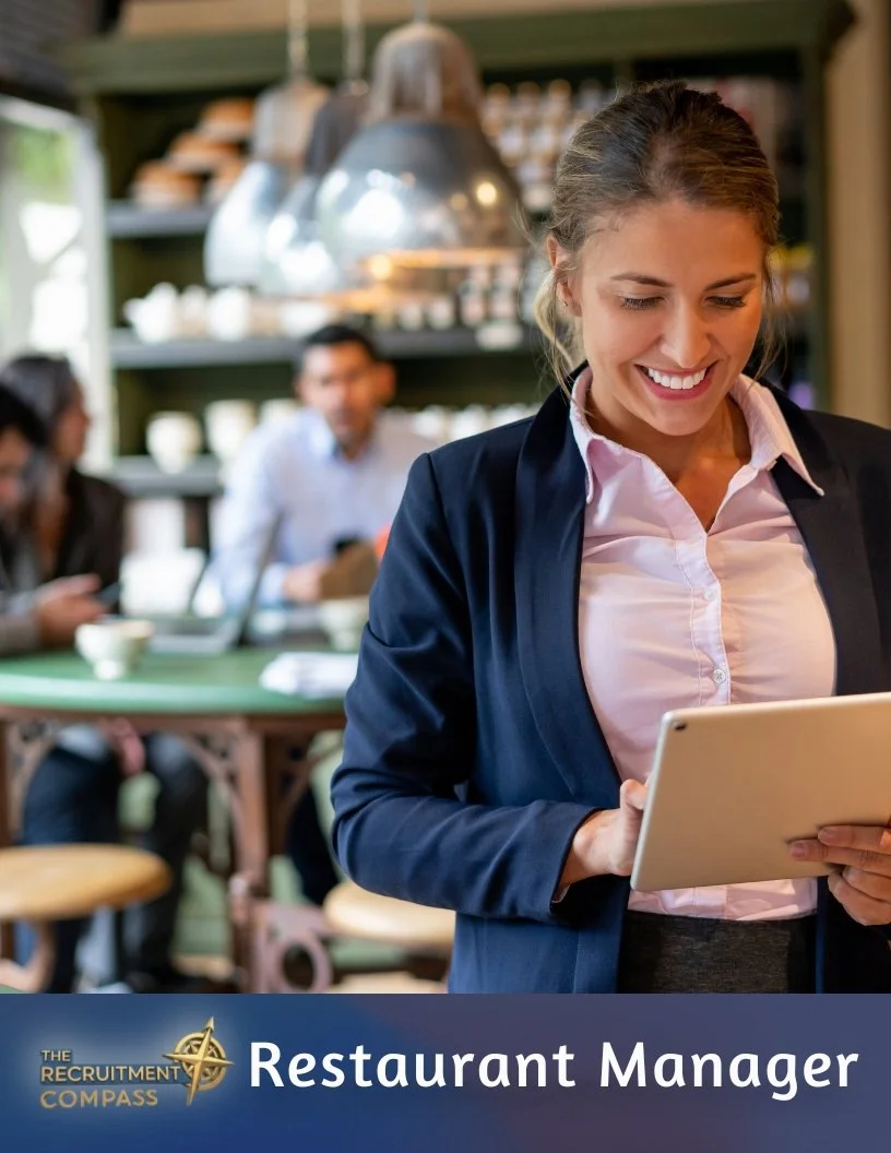 A smiling woman in business attire holding a tablet inside a restaurant, with other patrons and kitchen shelving in the background.