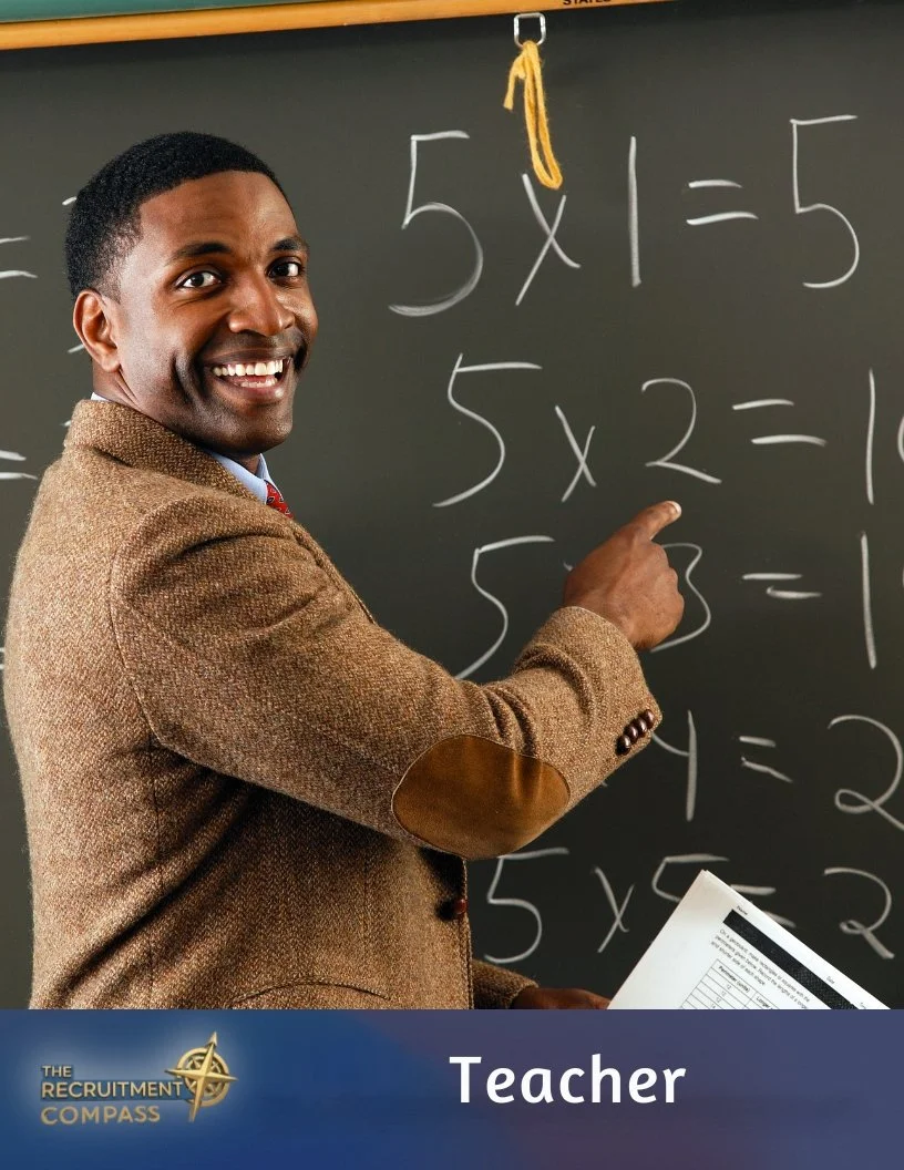 A smiling male teacher in a brown blazer points at a math problem on a blackboard with equations involving multiplication. He holds a sheet of paper in his other hand.