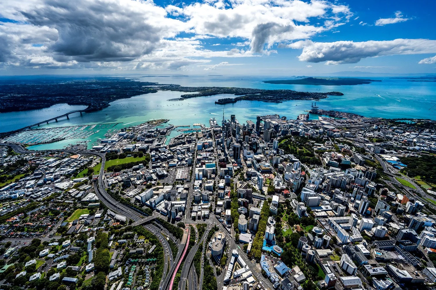 Aerial view of downtown Auckland, New Zealand, with high-rise buildings, urban streets, and surrounding water bodies, under a cloudy sky.