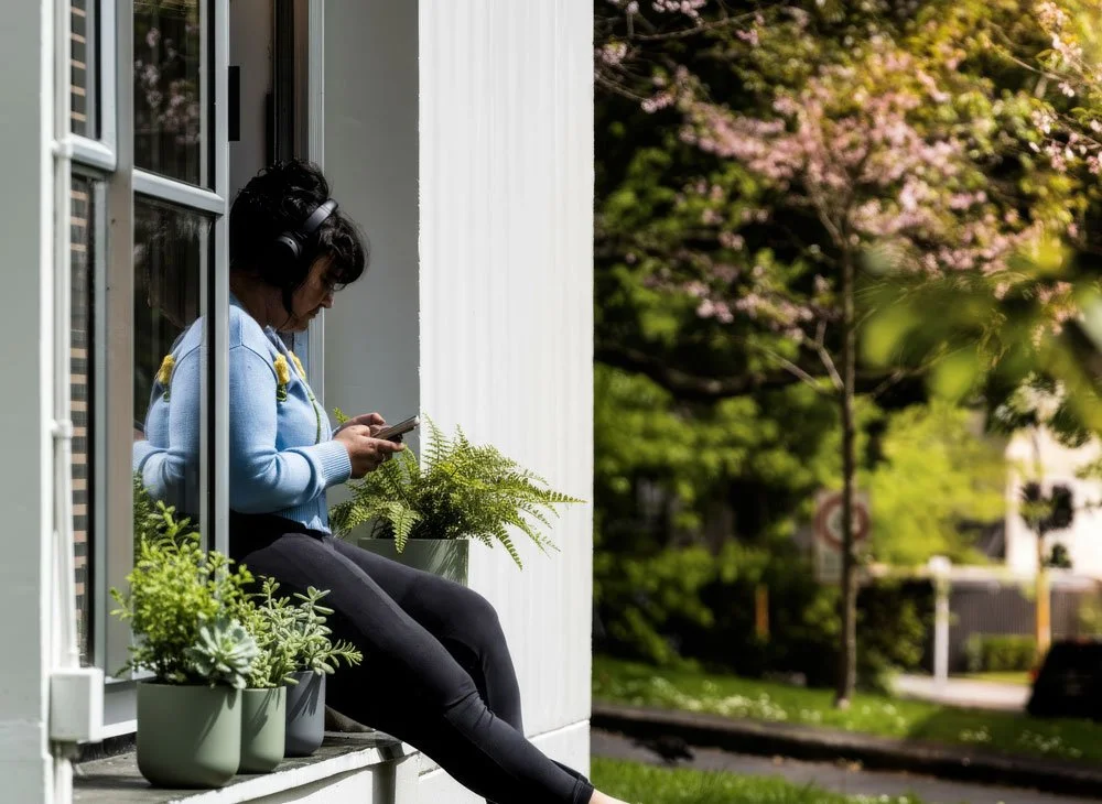 Woman sitting on windowsill wearing headphones, looking at her phone, with potted plants nearby, outdoors with trees and pink blossoms in the background.