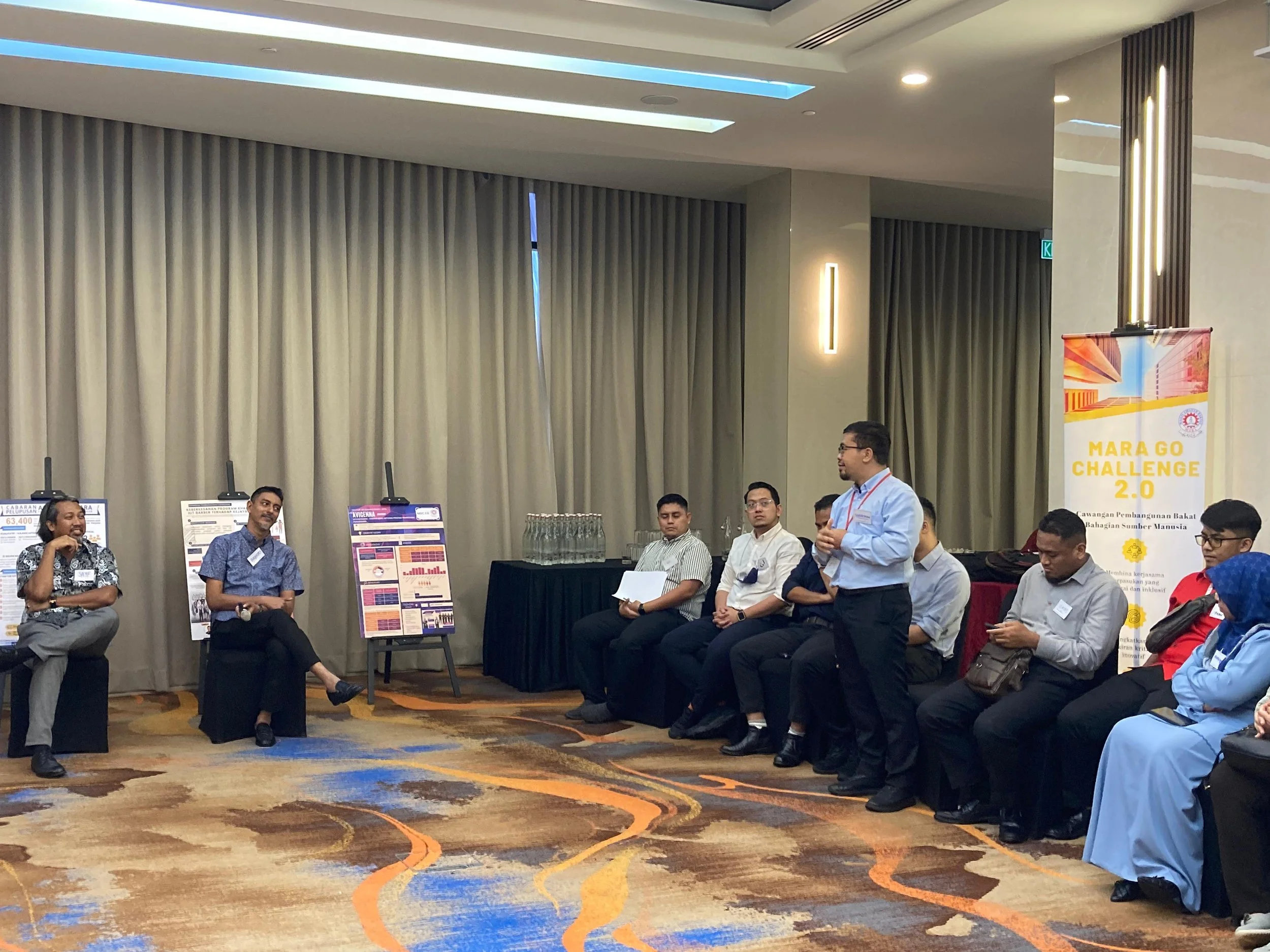A group of people sitting and standing in a conference room, with posters on easels and a banner that reads "MARA GO CHALLENGE 2.0" in the background.