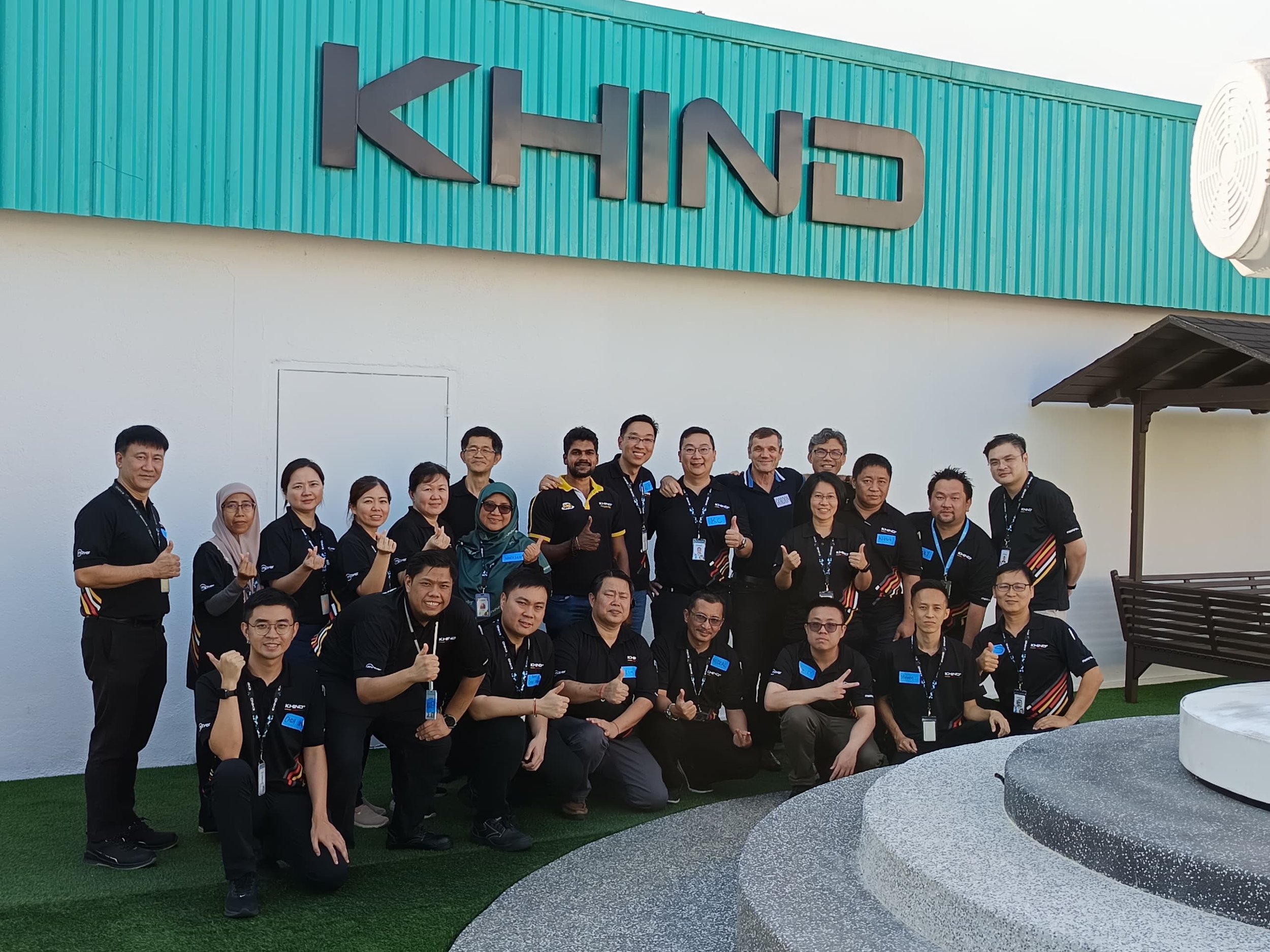 Group of people posing together outside a building with a sign reading "KIND". They are wearing matching black shirts with company logos and ID badges, some giving thumbs up.