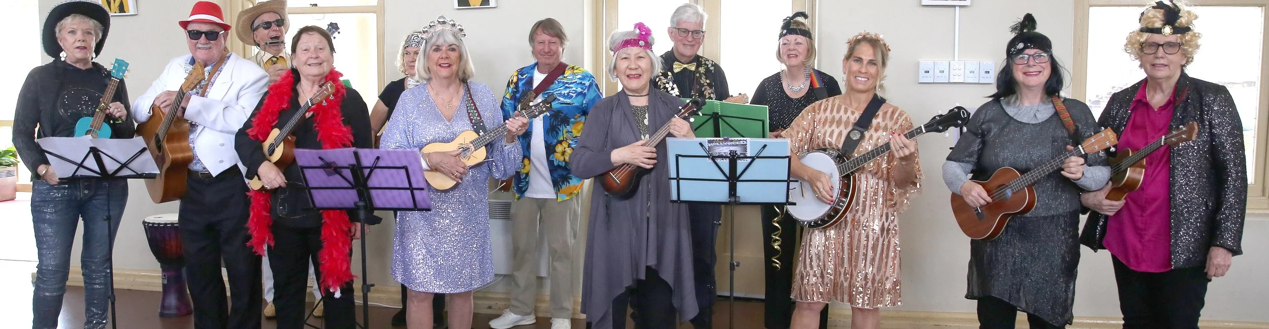 Six people performing with musical instruments at a gathering. Five are standing and playing guitars, one is sitting and playing drums. All are wearing purple hats, some with purple or black clothing, and are positioned in front of colorful artwork on the wall.