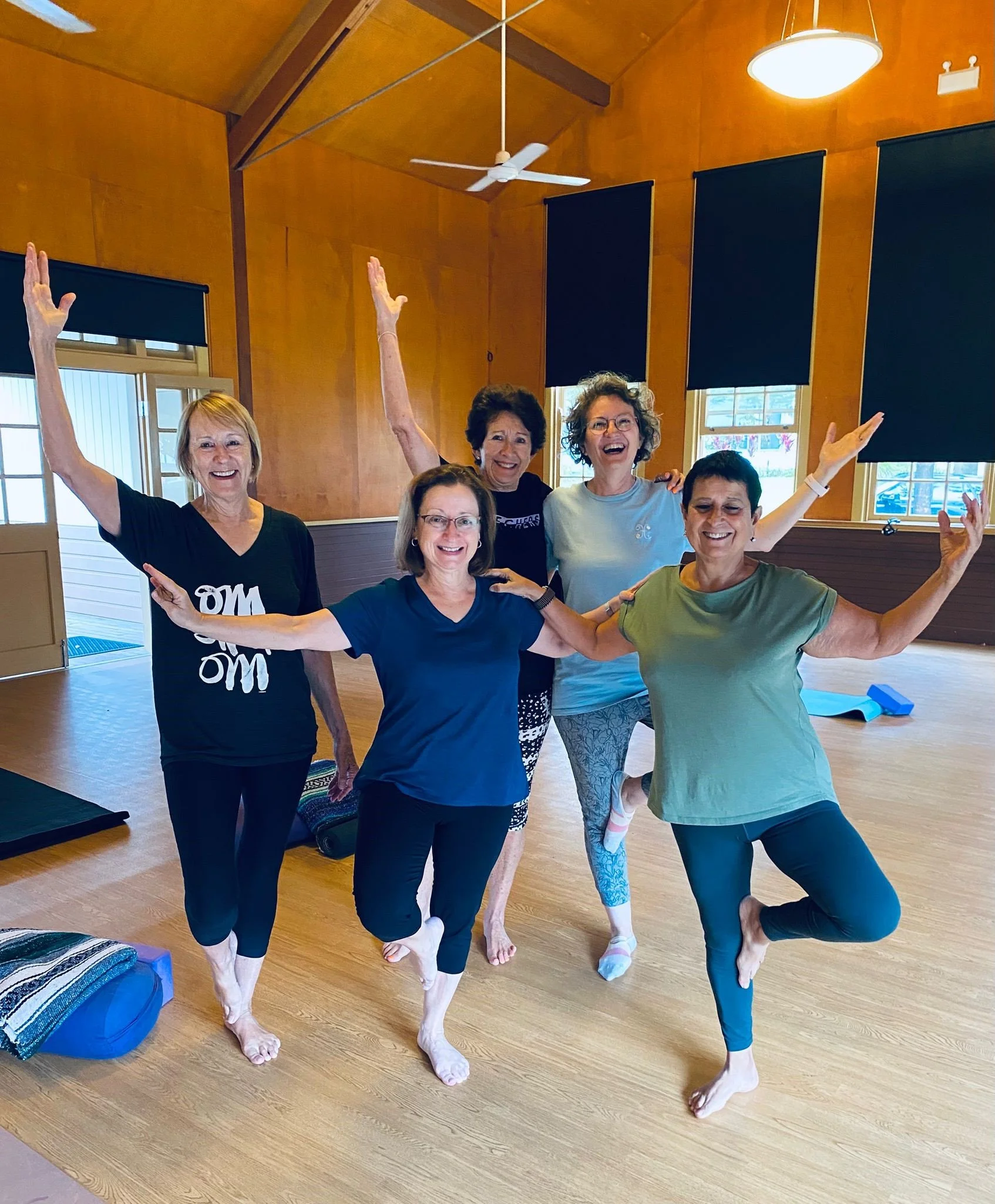 A group of people participating in a yoga class in a spacious, well-lit wooden room with tall windows and high ceilings. The instructor is at the front, demonstrating a pose, while students sit on mats with arms extended in a seated variation of a stretch.