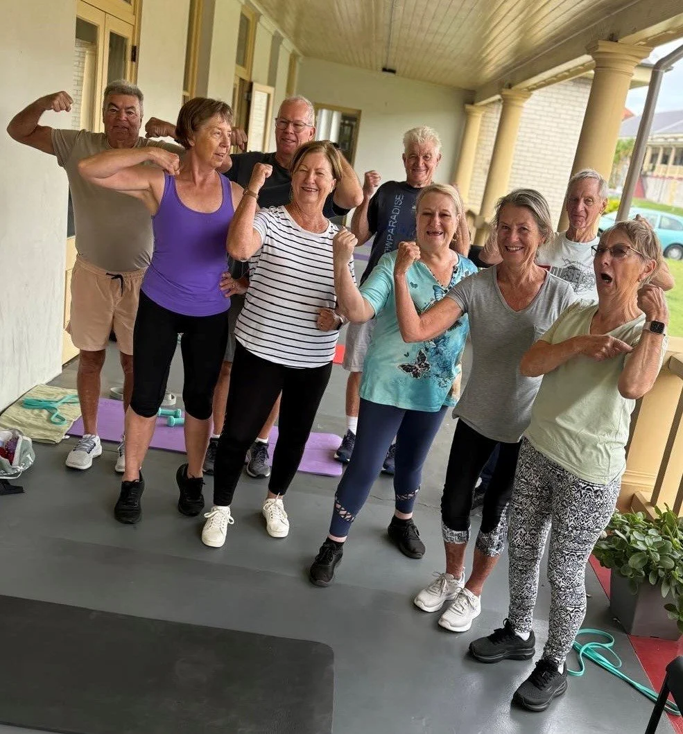 Older adults participating in a seated exercise class, standing and holding weights above their heads in a spacious room with paintings on the walls.