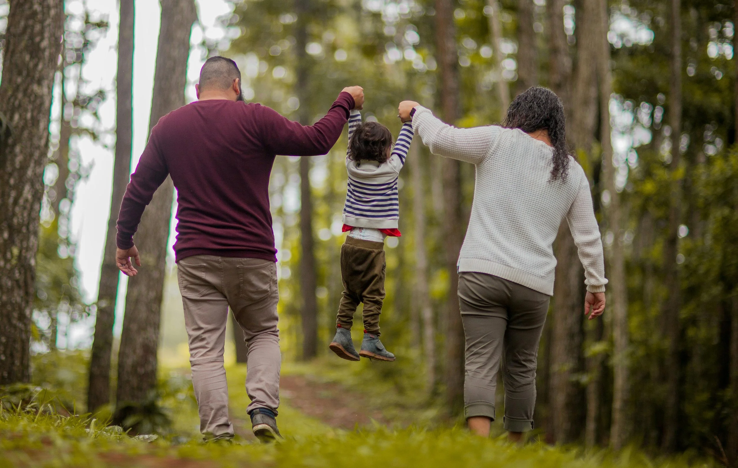 A family of three, two adults and a child, walking and playing in a forested area, with the child being lifted in the air by the adults.