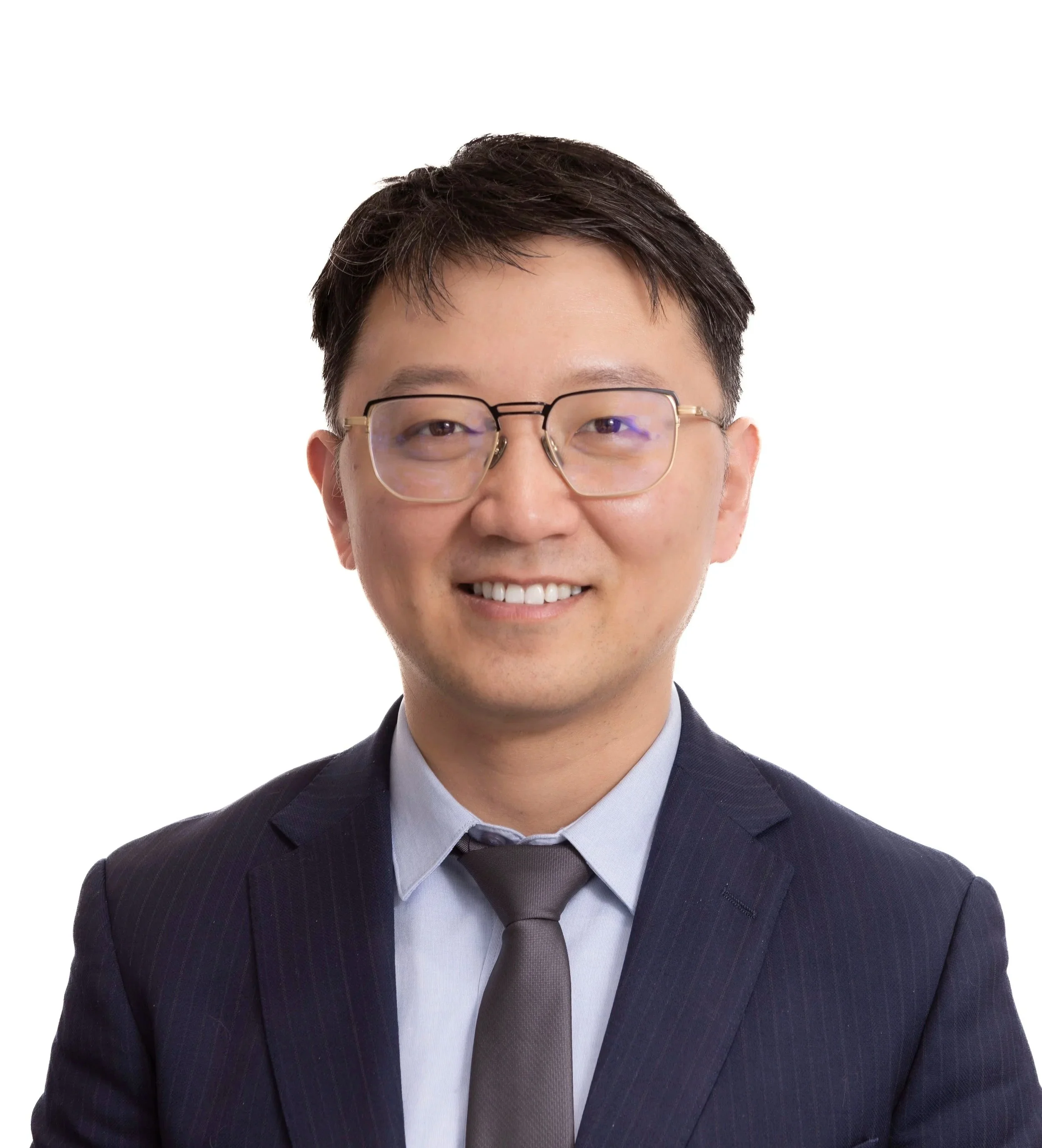 A smiling man in a dark suit, light blue shirt, and gray tie, wearing glasses, with short dark hair, set against a white background.
