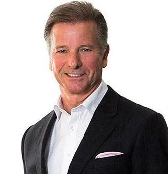 A middle-aged man with short, brown hair, wearing a dark suit jacket and a white dress shirt, smiling at the camera against a plain background.