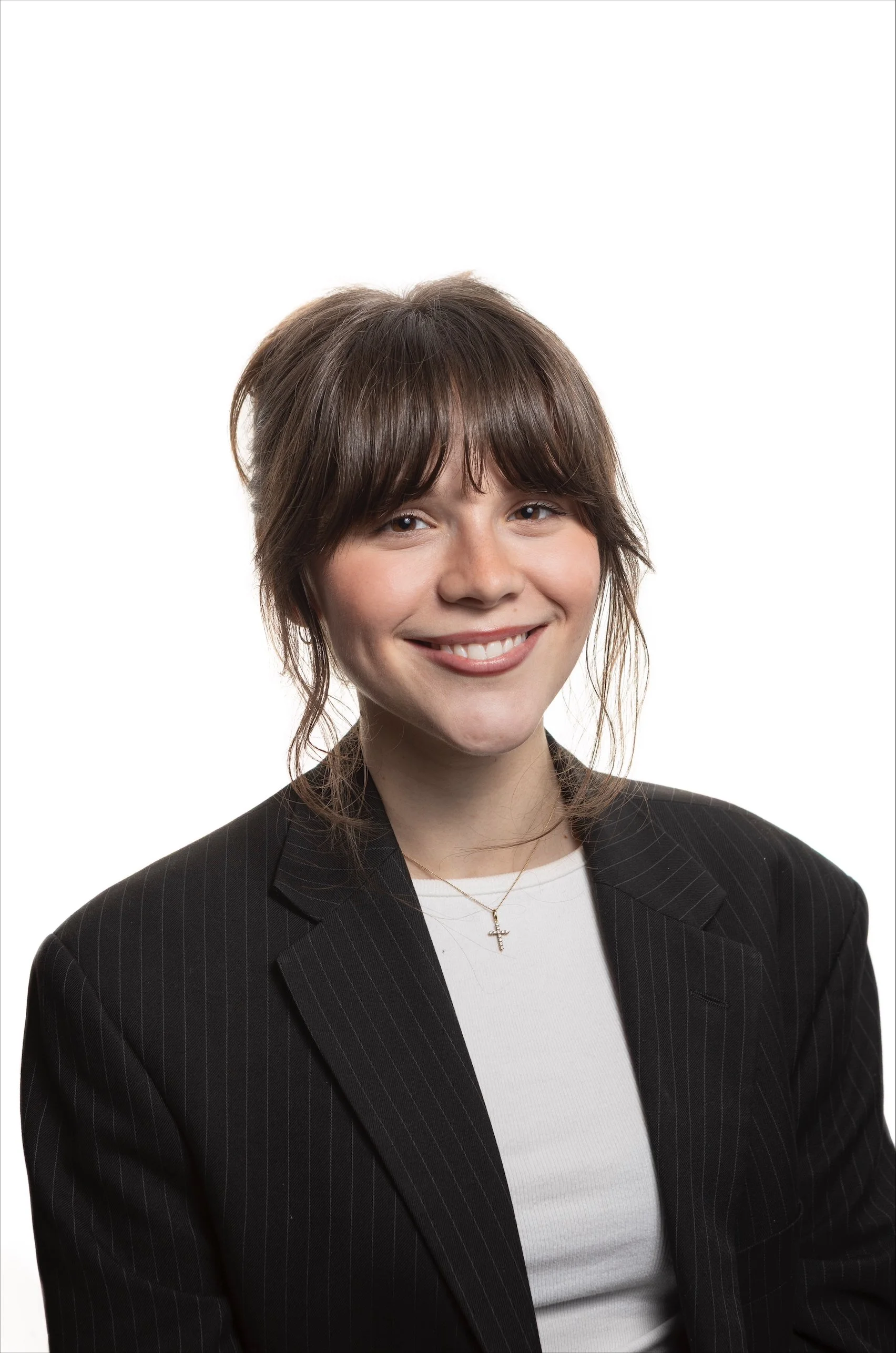 A woman with shoulder-length brown hair smiling, wearing a black pinstripe blazer, a white shirt, and a gold necklace with a cross pendant, posed against a plain white background.