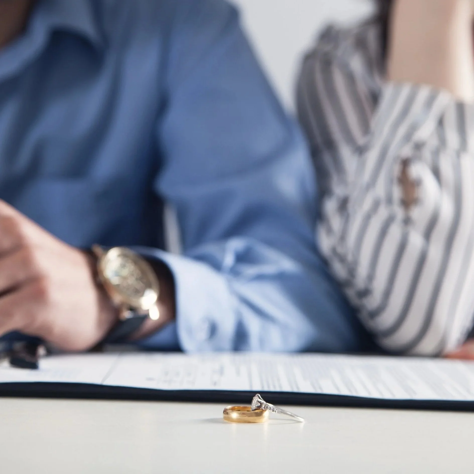 Close-up of wedding rings on a table with a blurred blue-shirted man and striped shirt woman in the background.