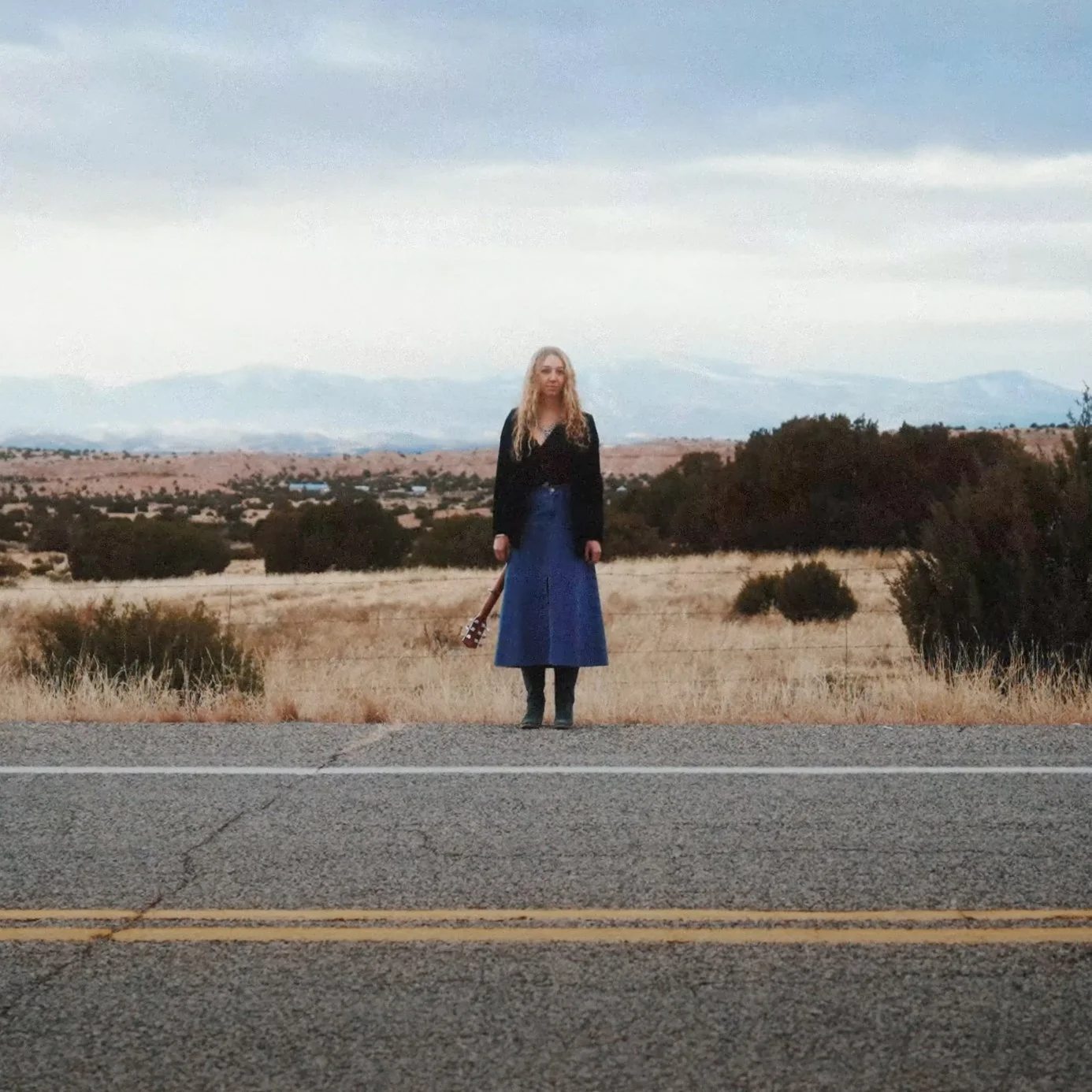 A woman stands on the side of a road in a rural area, holding a guitar, with a landscape of open fields, bushes, and distant mountains under a cloudy sky.