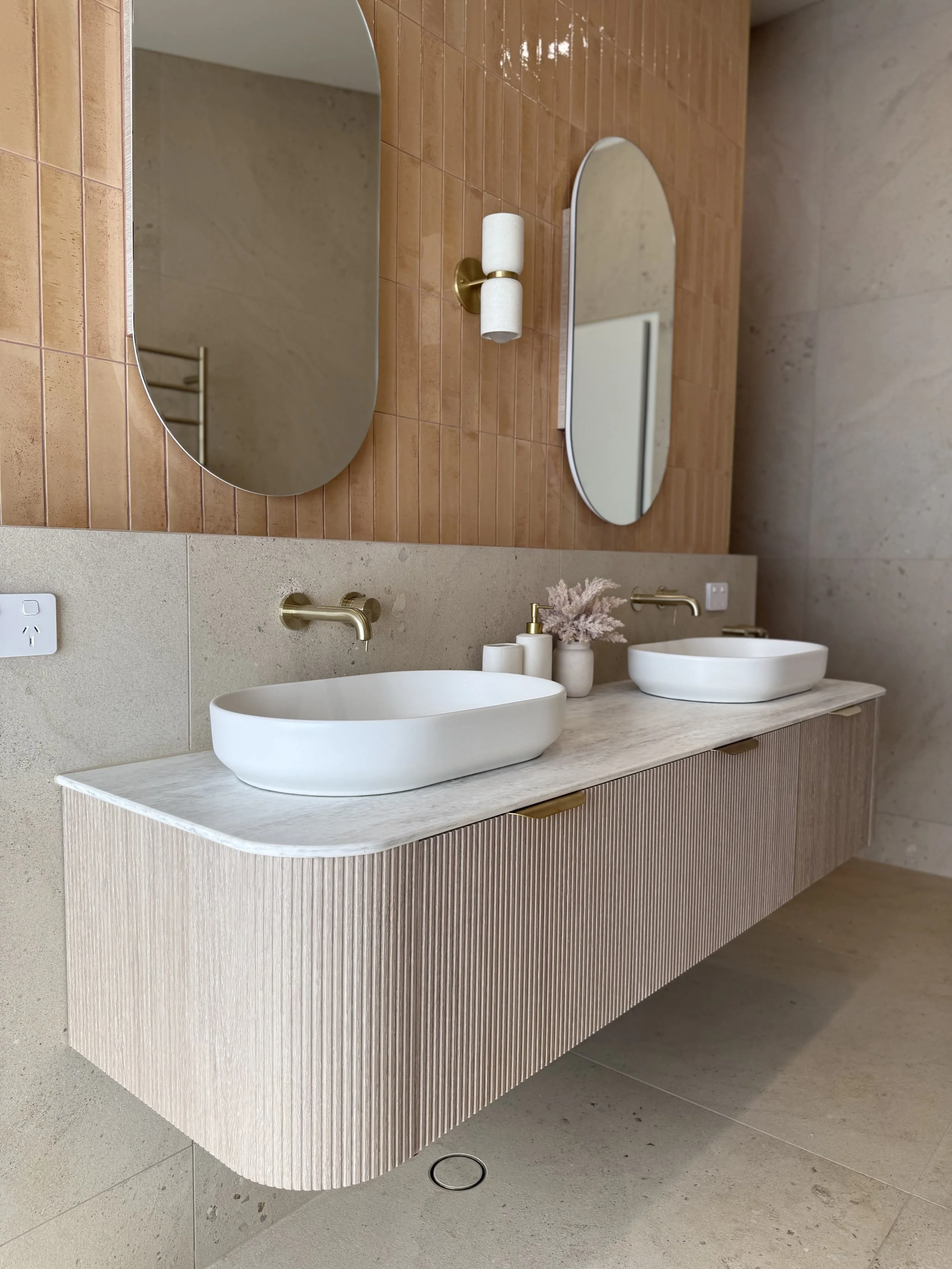 Double bathroom vanity with two white vessel sinks, gold wall-mounted faucets, two oval mirrors above each sink, and a beige cabinet with vertical grooves. Decor includes a soap dispenser, two small vases, and a wall sconce with two white cylindrical shades. The wall behind the vanity features orange-brown tiles, and the surrounding wall is a light beige color. There's a beige countertop and a visible electrical outlet on the wall.