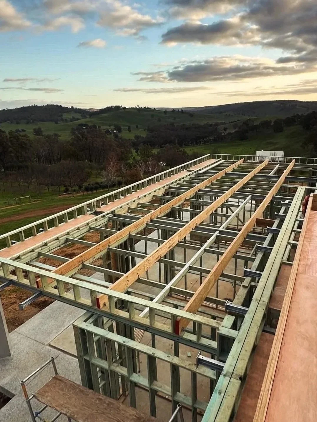 Construction of a building's wooden framework with a scenic rural landscape of rolling hills and scattered trees in the background.