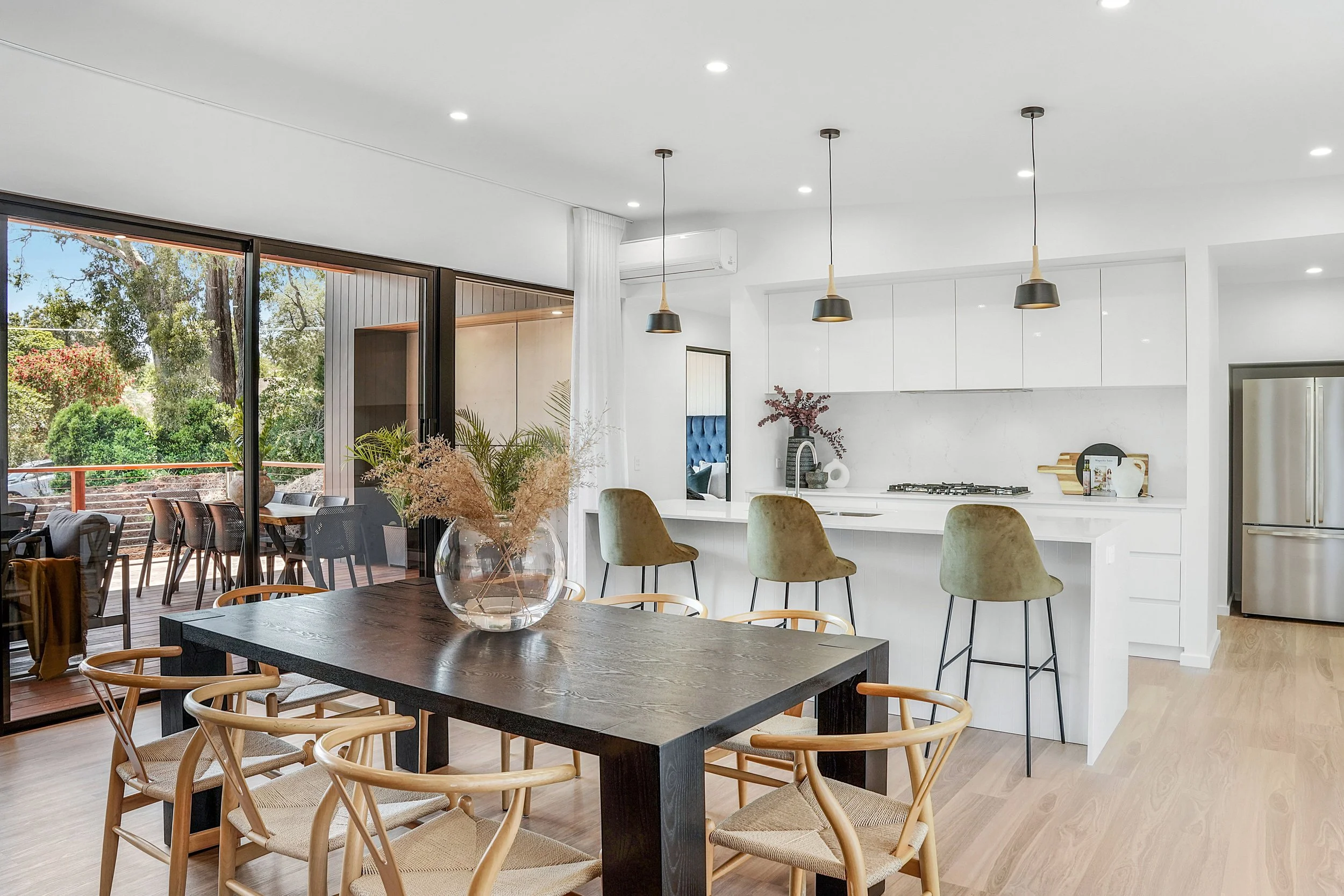 Interior of a modern, open-concept kitchen and dining area with a large window, wooden dining table, tan chairs, white cabinetry, pendant lighting, and outdoor patio view.