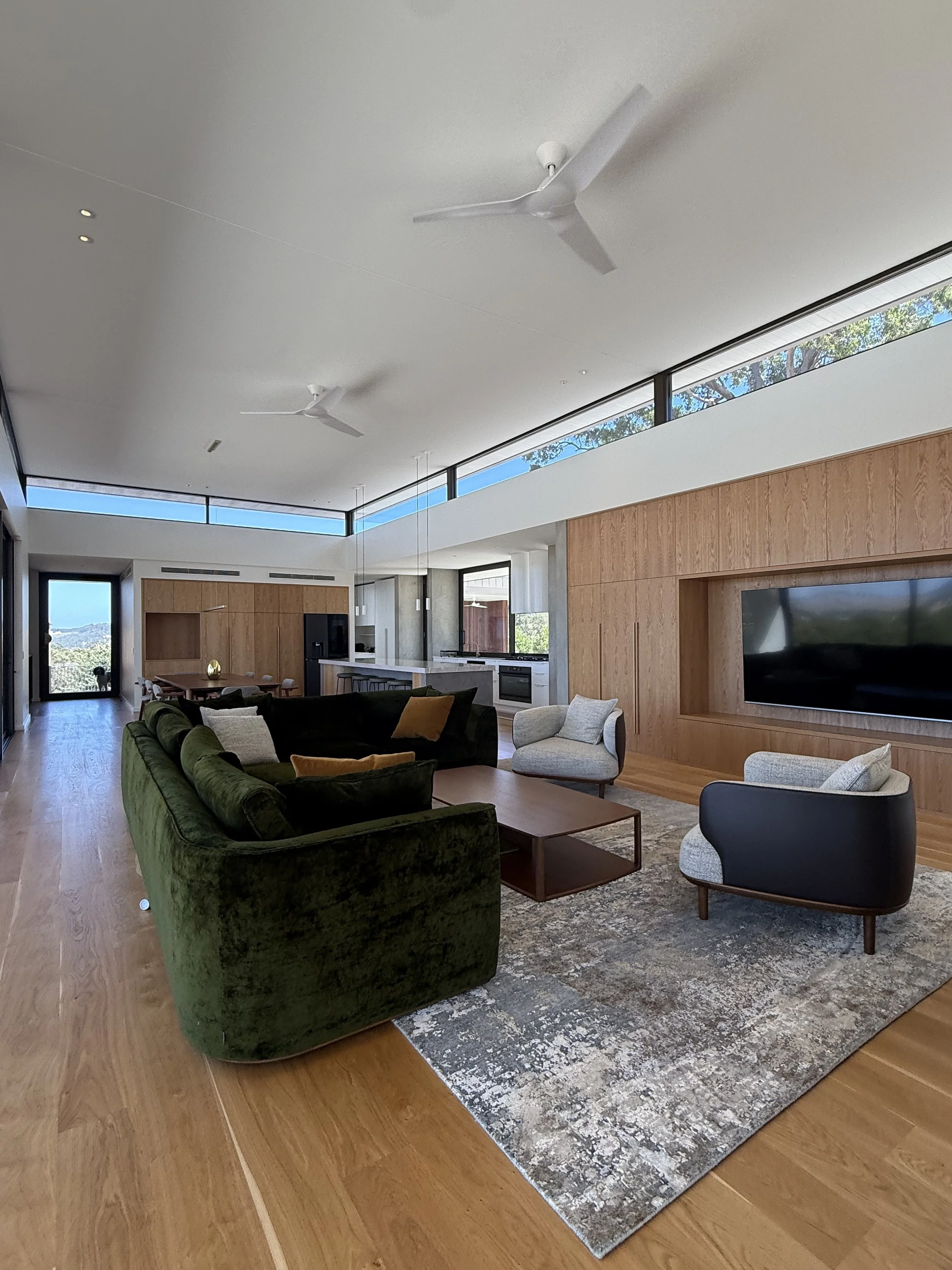 Open-plan living room with green velvet sofa, white and black armchairs, a rug, wooden flooring, a large TV on wooden wall panel, and a view of trees outside through high windows.