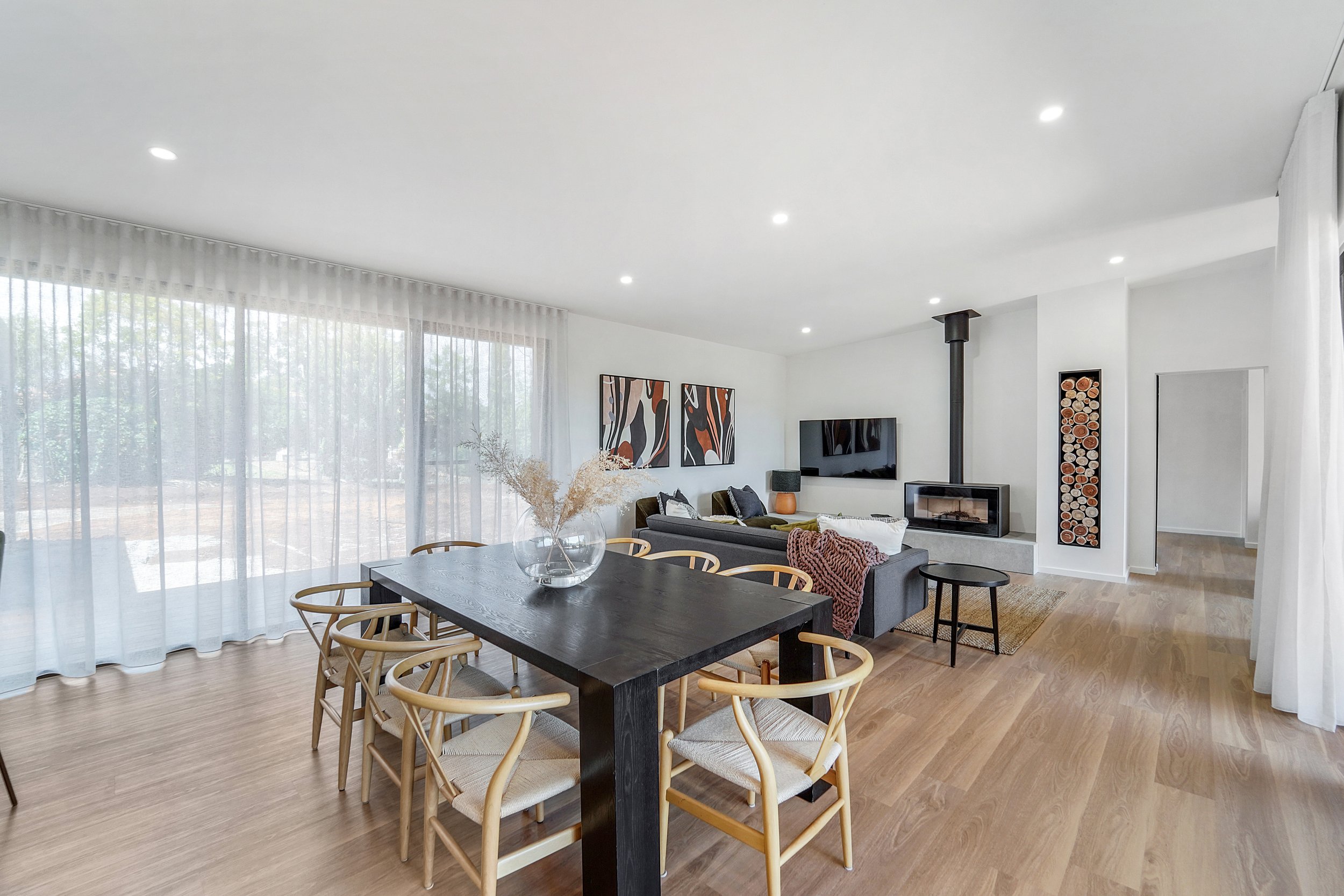 A modern living and dining room with large windows covered by sheer white curtains, wooden flooring, a black dining table with wicker chairs, a gray sofa, a TV mounted on the wall, colorful abstract artwork, and a wood-burning stove.