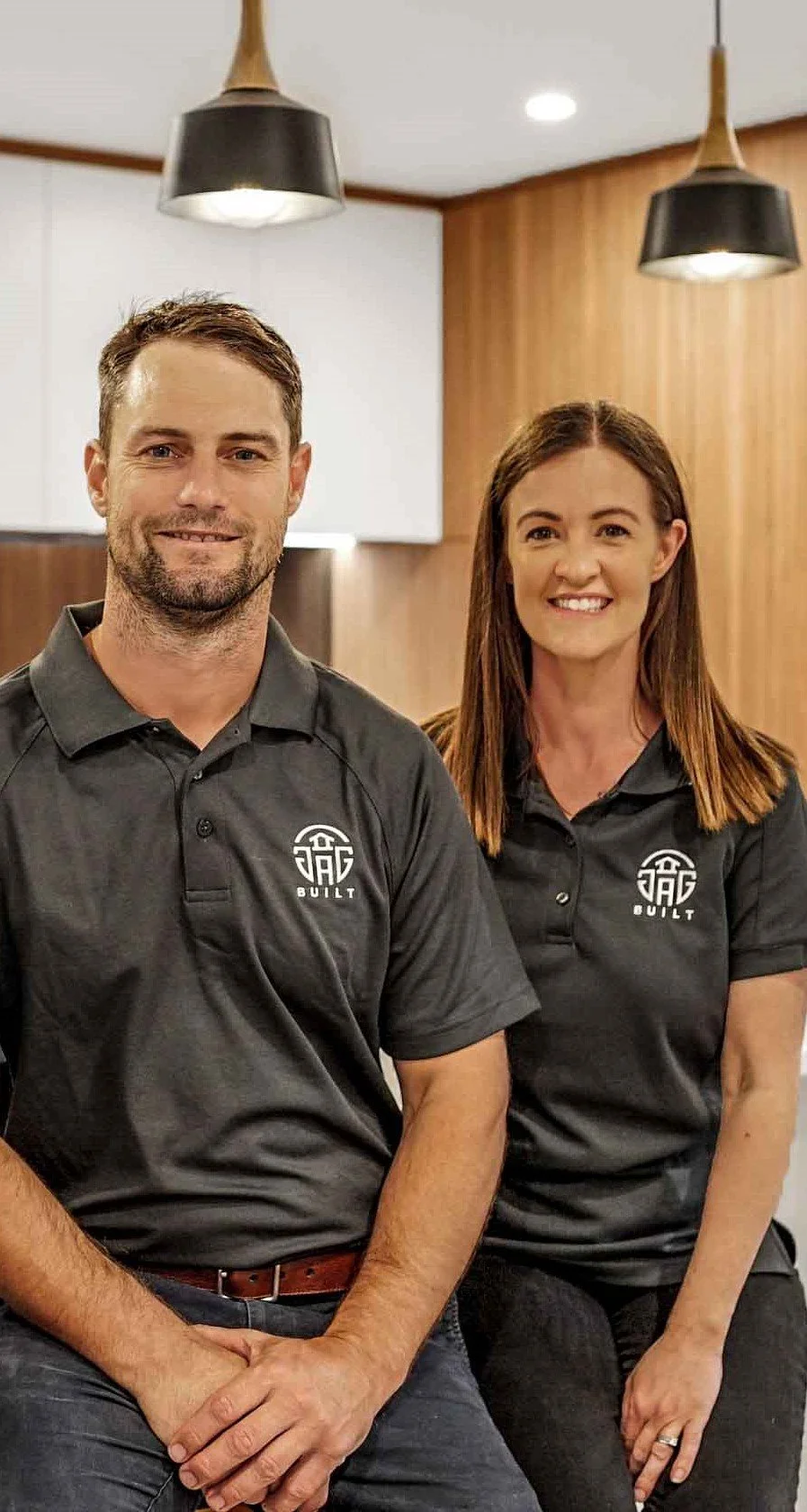 Two people, a man and a woman, wearing matching black polo shirts with a logo that reads "Built" pose for a photo inside a modern, well-lit room with wooden accents and hanging ceiling lamps.
