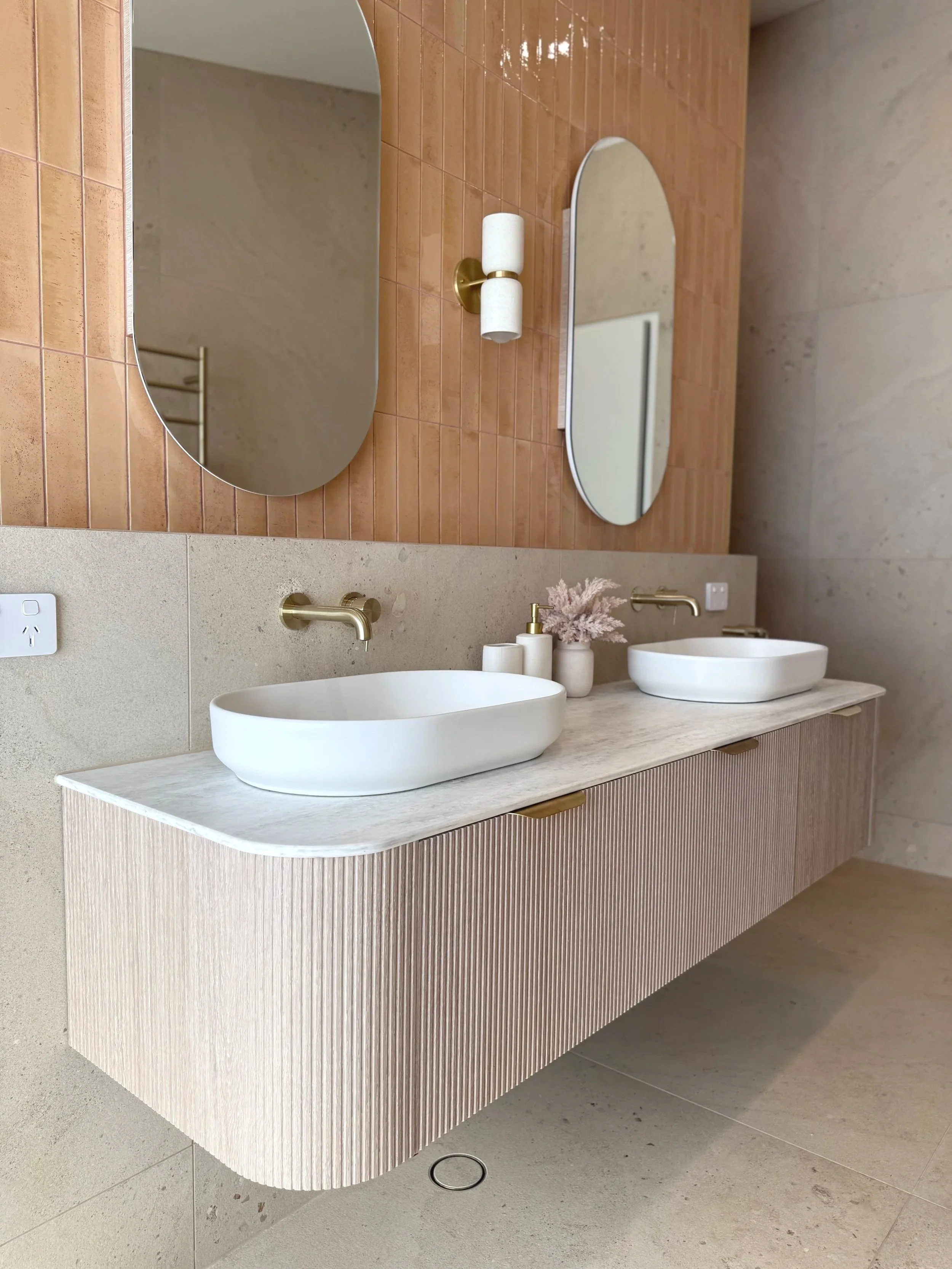 Modern bathroom with a dual oval mirror vanity, two white vessel sinks, gold fixtures, beige textured cabinetry, and decorative vases with pink dried flowers on a marble countertop.