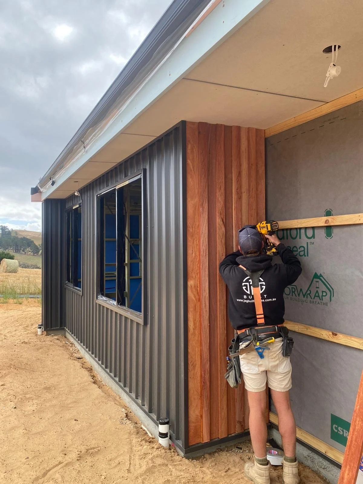 A construction worker is installing wooden panels on the exterior wall of a building under construction. The building has metal siding and windows, and the worker is using a power drill. The scene is outdoors on a cloudy day.