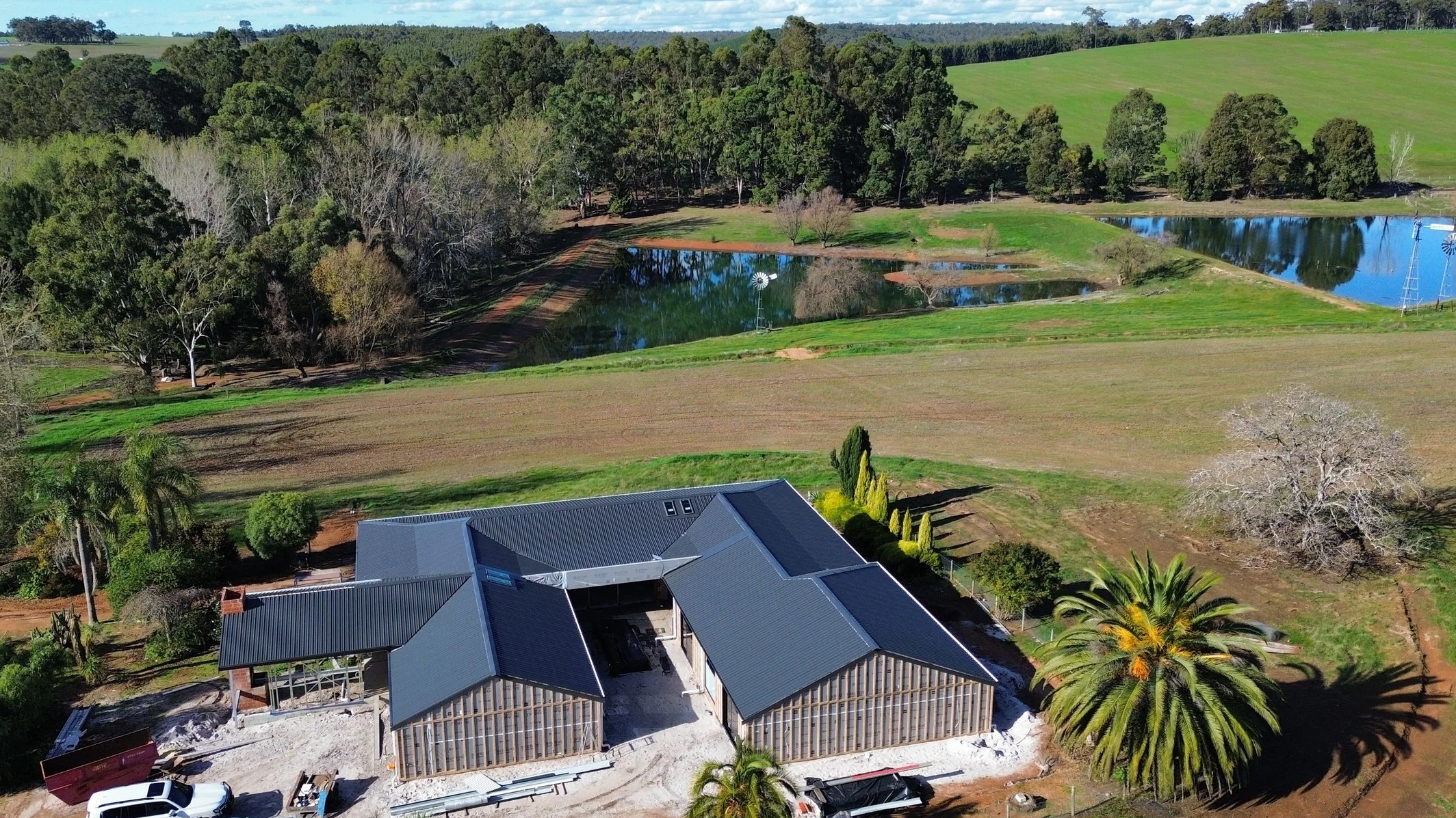 Aerial view of a house under construction with a gray metal roof, surrounded by green trees, fields, and a pond, with wind turbines in the background.