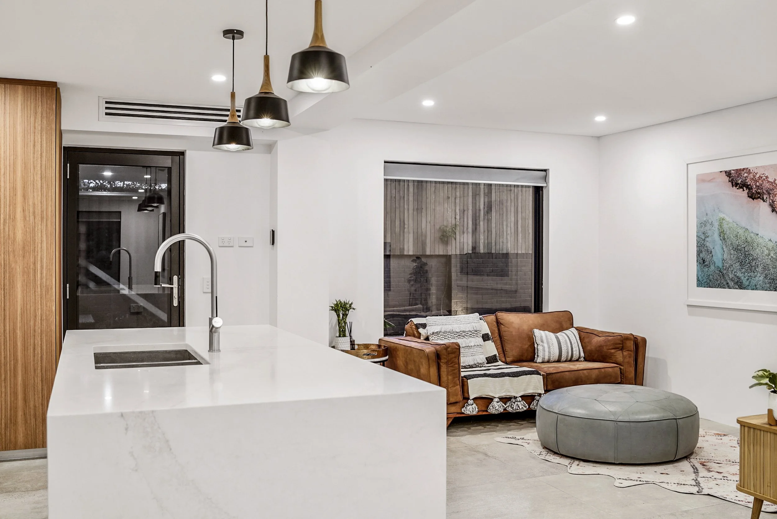 Modern living room with white walls, a leather sofa with striped and plain pillows, a round gray ottoman, a colorful abstract painting, and large windows showing an outdoor area with wooden fencing.