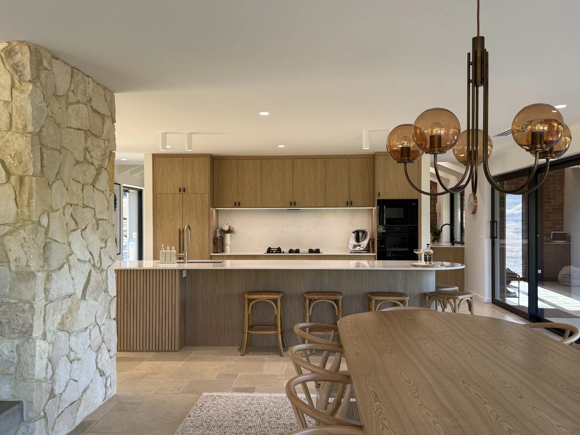 Modern kitchen with wooden cabinets, stone wall, pendant light fixtures, kitchen island with bar stools, and a dining table with chairs.