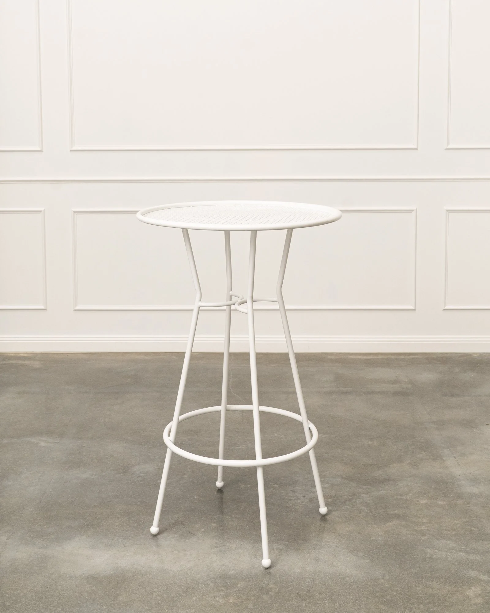 A white metal bistro table with a round mesh top and three thin legs, standing on a concrete floor in front of white paneled walls.
