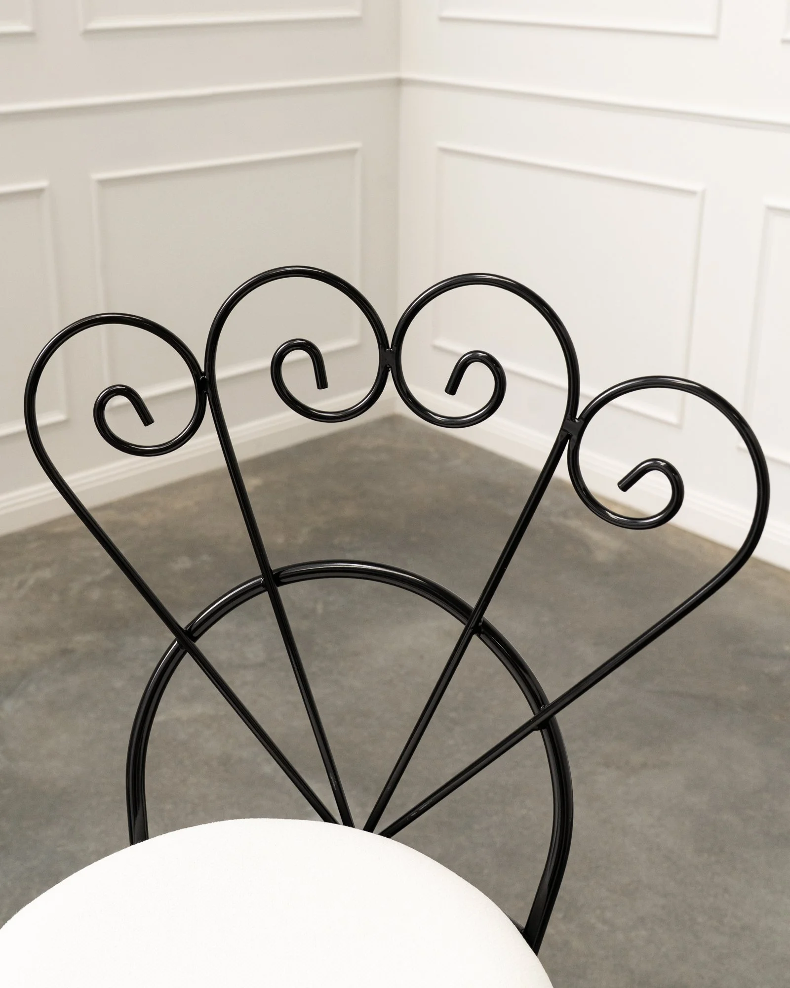 Close-up of a black metal chair with a pad cushion and a backrest featuring a decorative scroll design, in a room with white paneled walls and gray flooring.