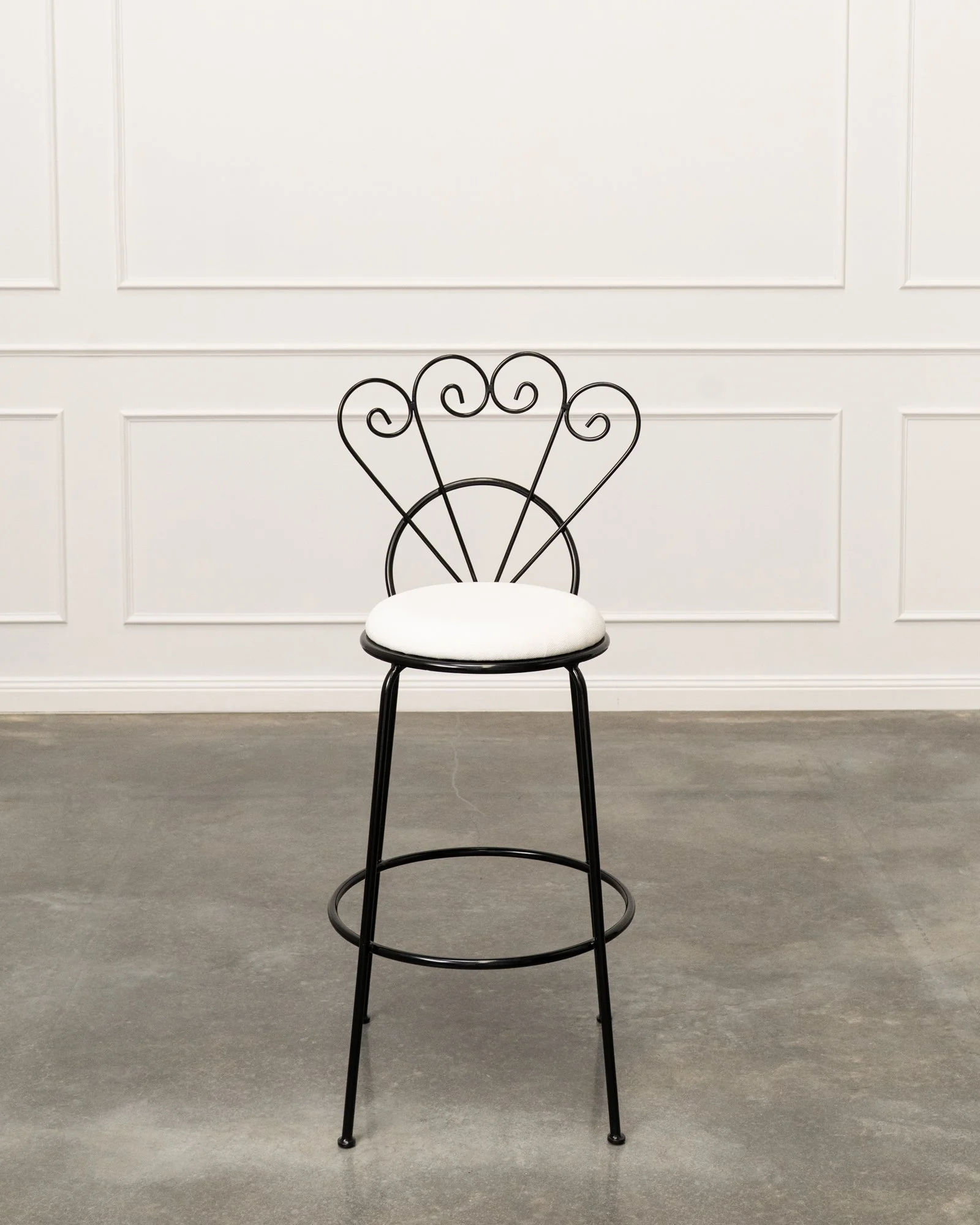 Black decorative metal chair with a white cushioned seat against a white paneled wall and gray floor.