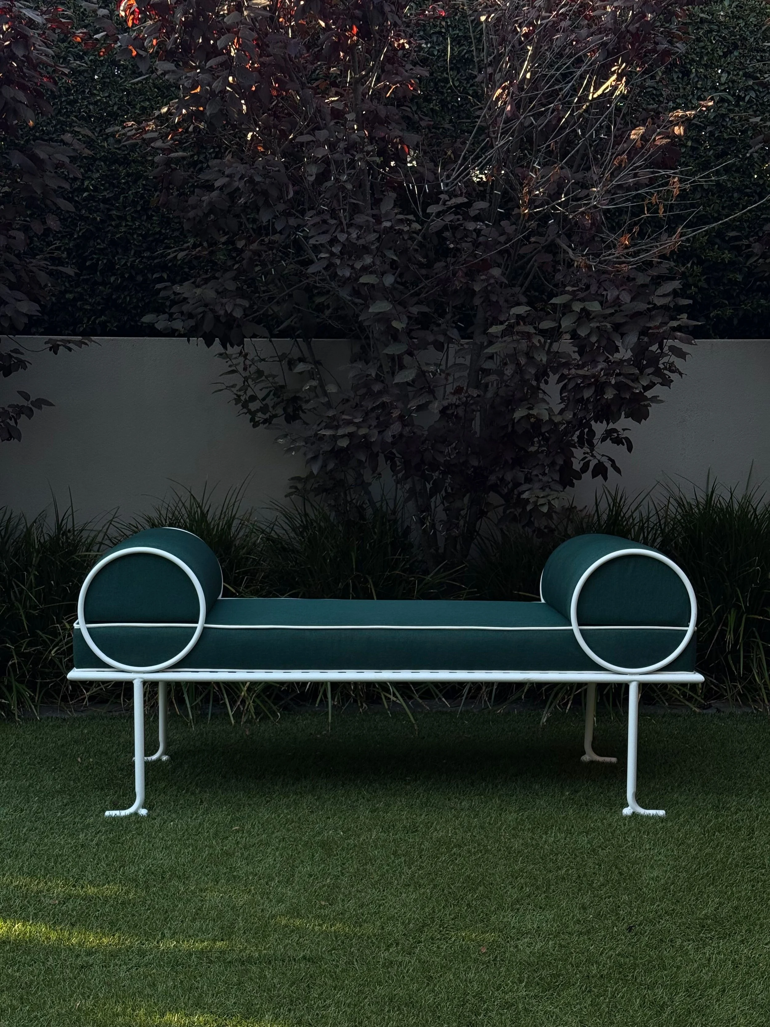 A modern, green bench with white metal frame, situated on a grassy area in front of dark foliage and trees.