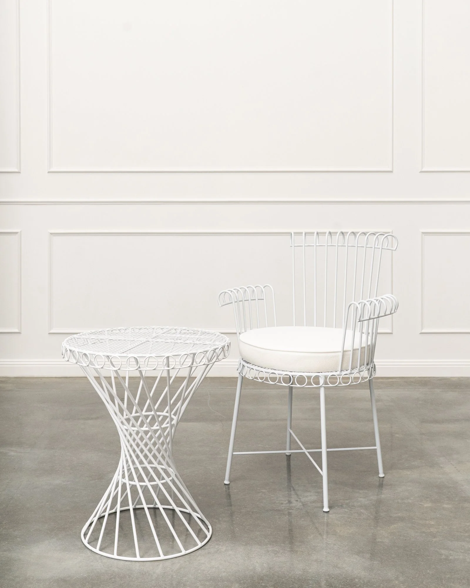 White wireframe round table and matching white wireframe chair with cushioned seat against white paneled wall with gray wood floor.