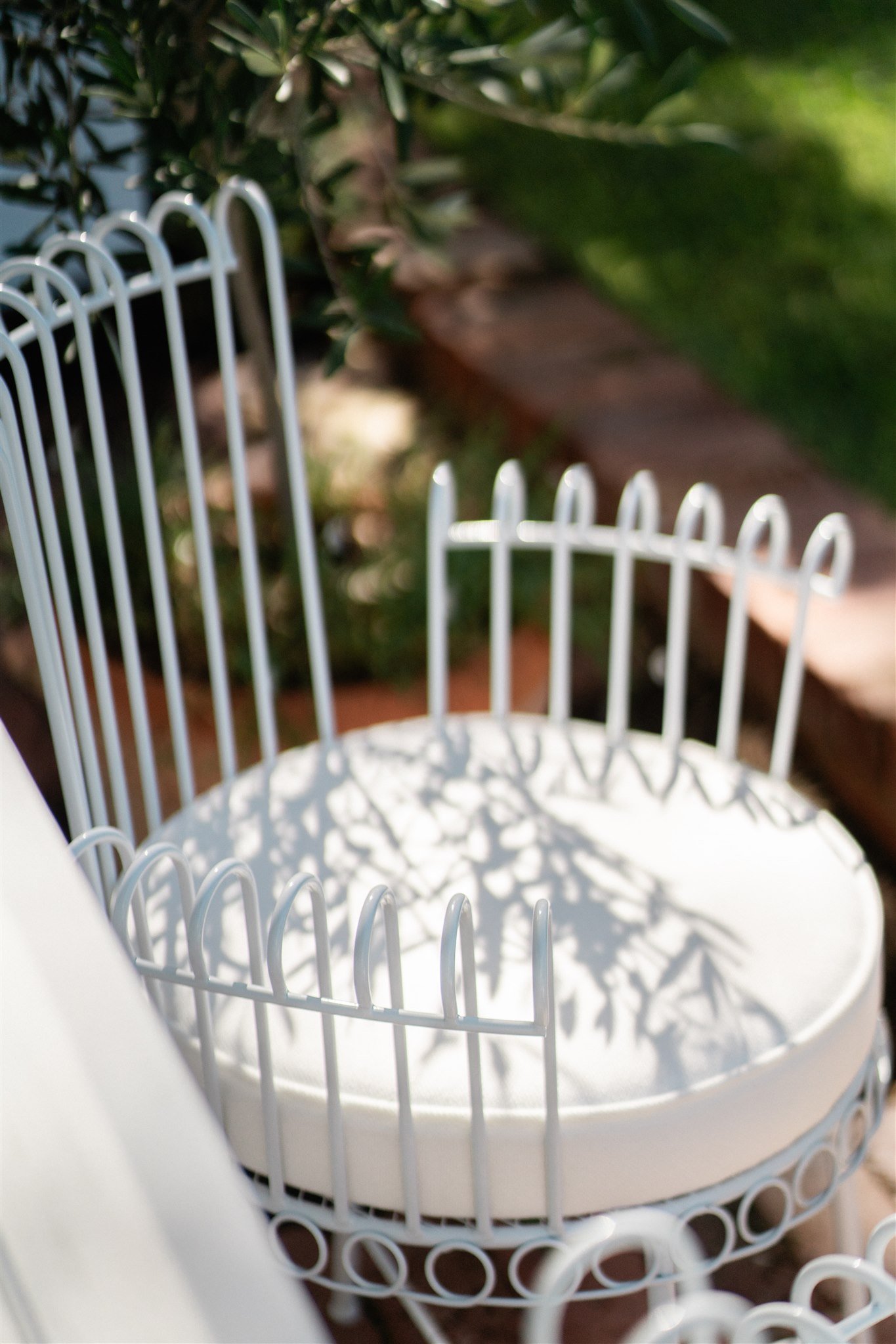 Close-up of a white wire chair with curved backrest cast shadows on the seat, outdoors with green foliage in the background.