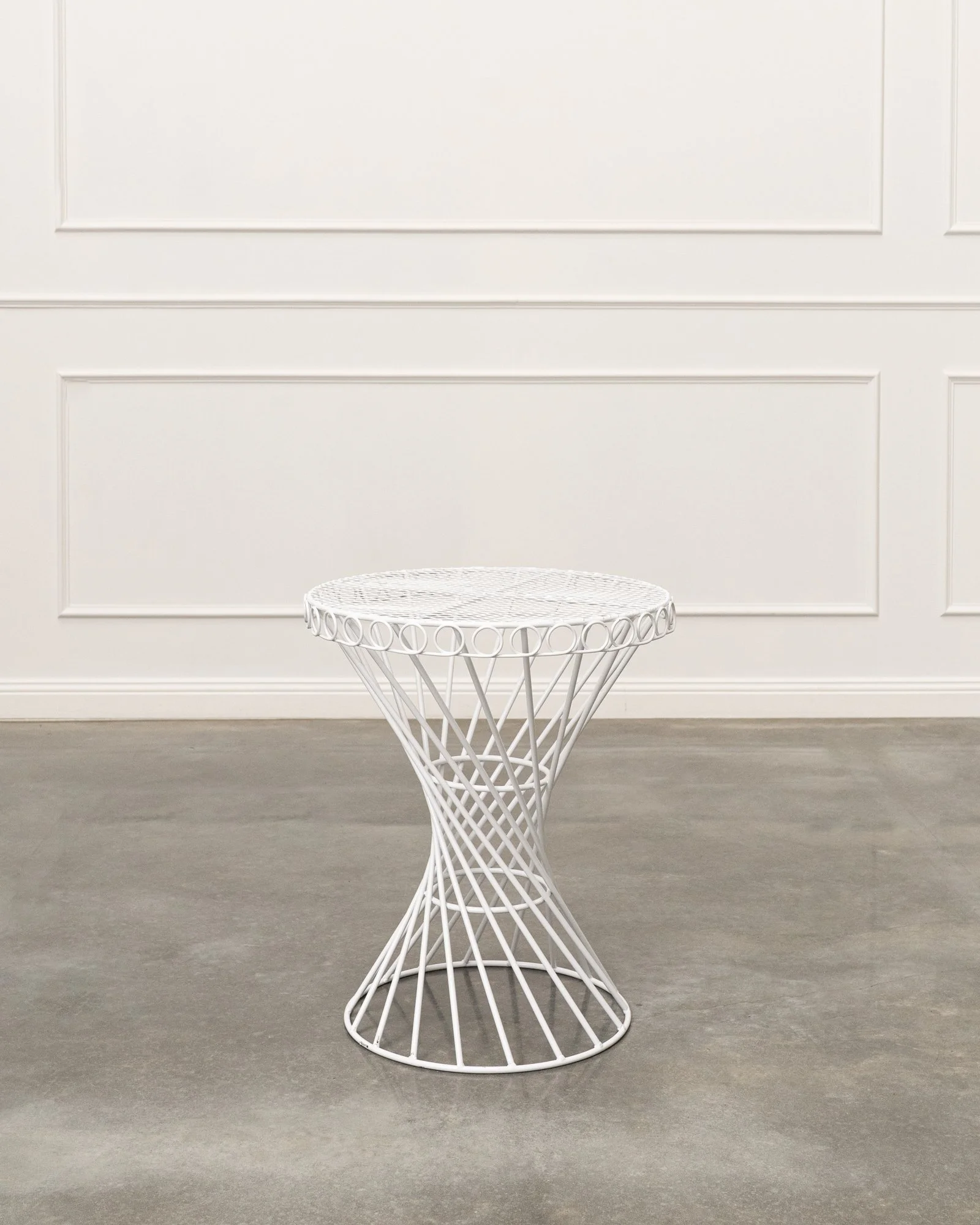 White round wire side table in front of plain white paneled wall with gray flooring.