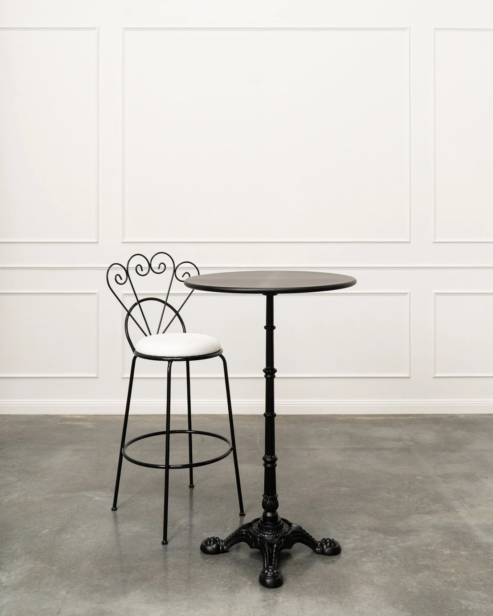 A black round table with four curved legs and a decorative pedestal base, accompanied by a metal chair with a white cushioned seat and an intricate backrest design, set against a white wall with decorative molding and a concrete floor.