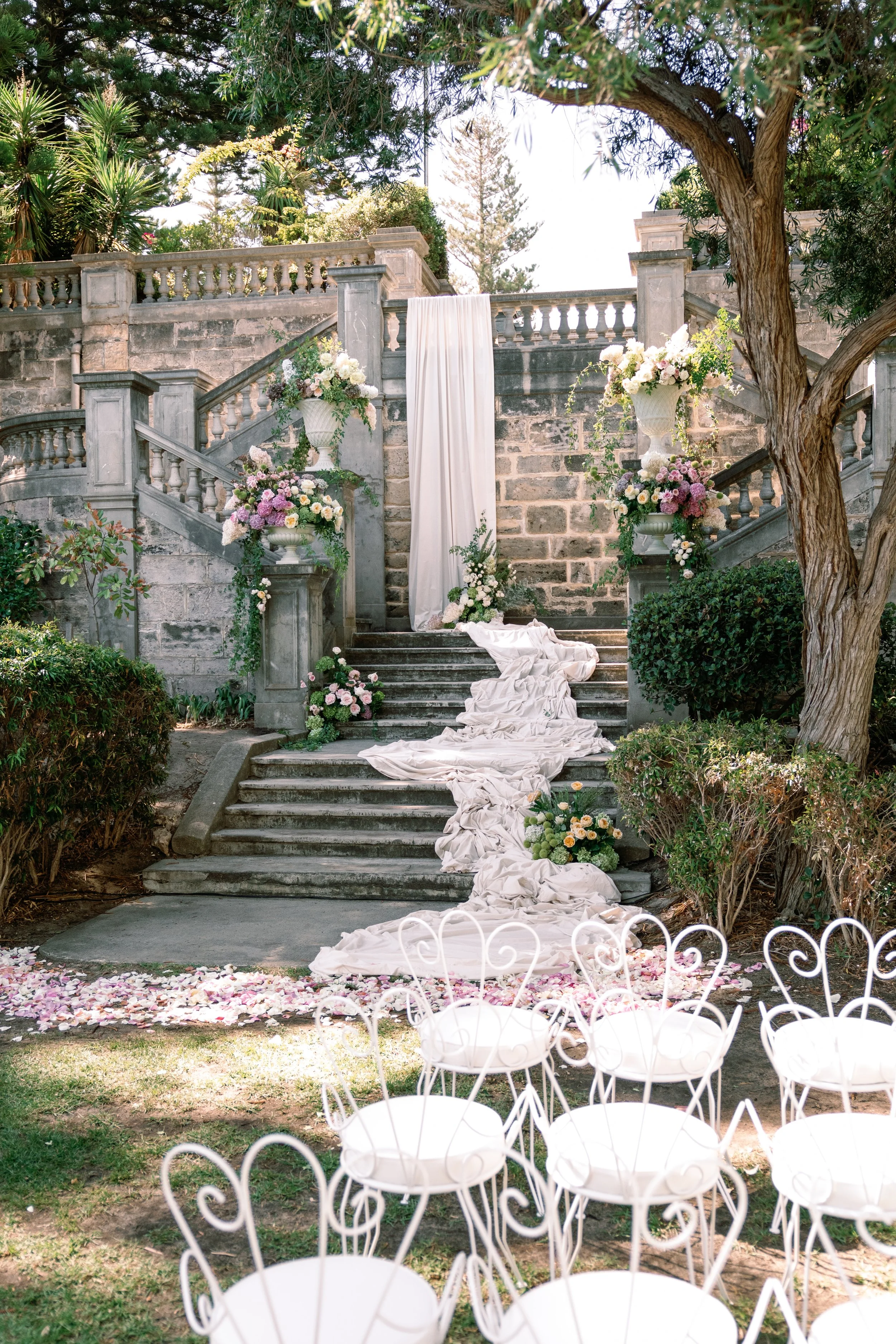 A stone staircase decorated with floral arrangements leading up to a draped white fabric backdrop, set in a garden with trees and chairs arranged in a semicircle in the foreground, suggesting a wedding or outdoor event setting.
