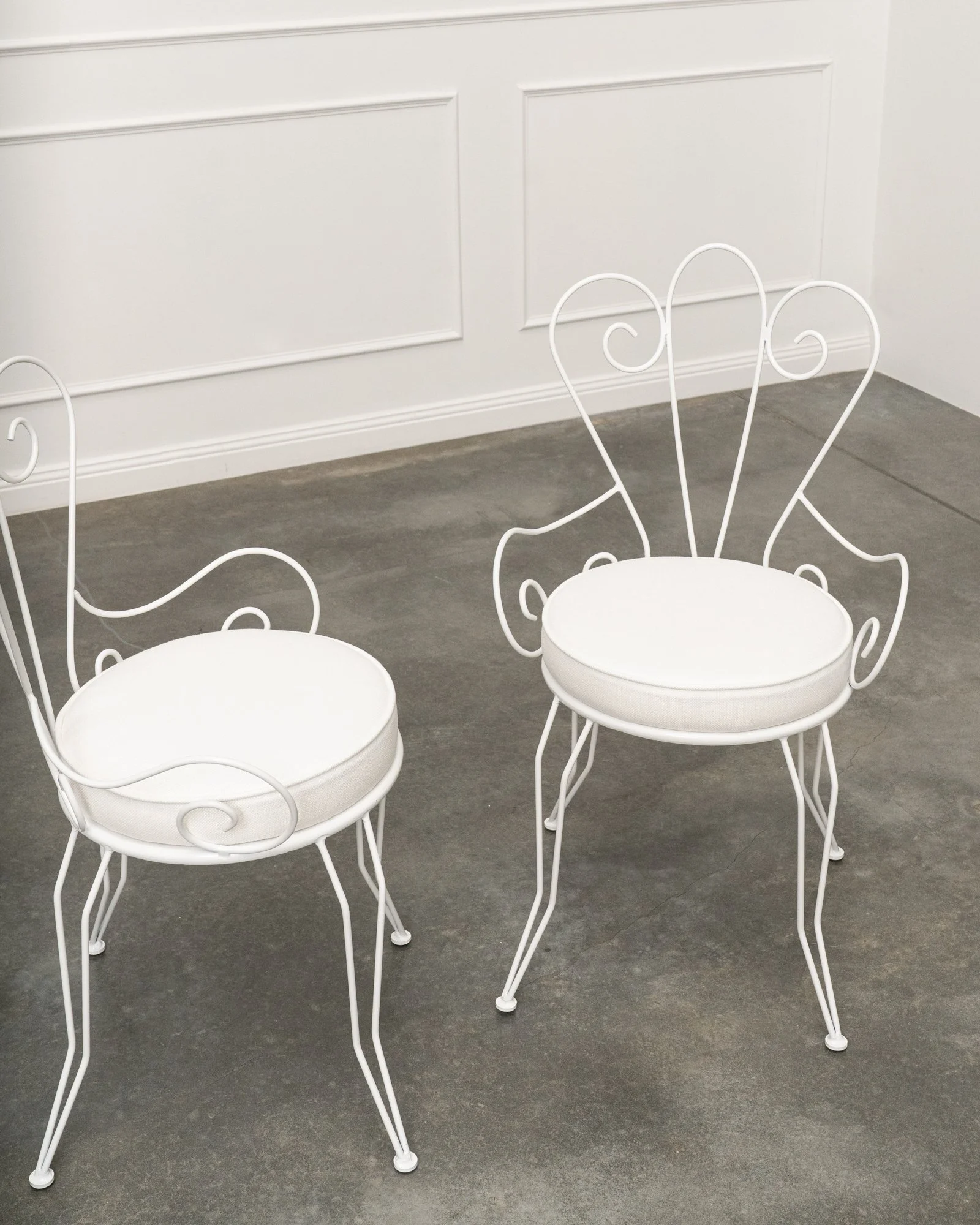White metal chairs with curved decorative backs in an indoor setting with white wainscoting wall and gray concrete floor.
