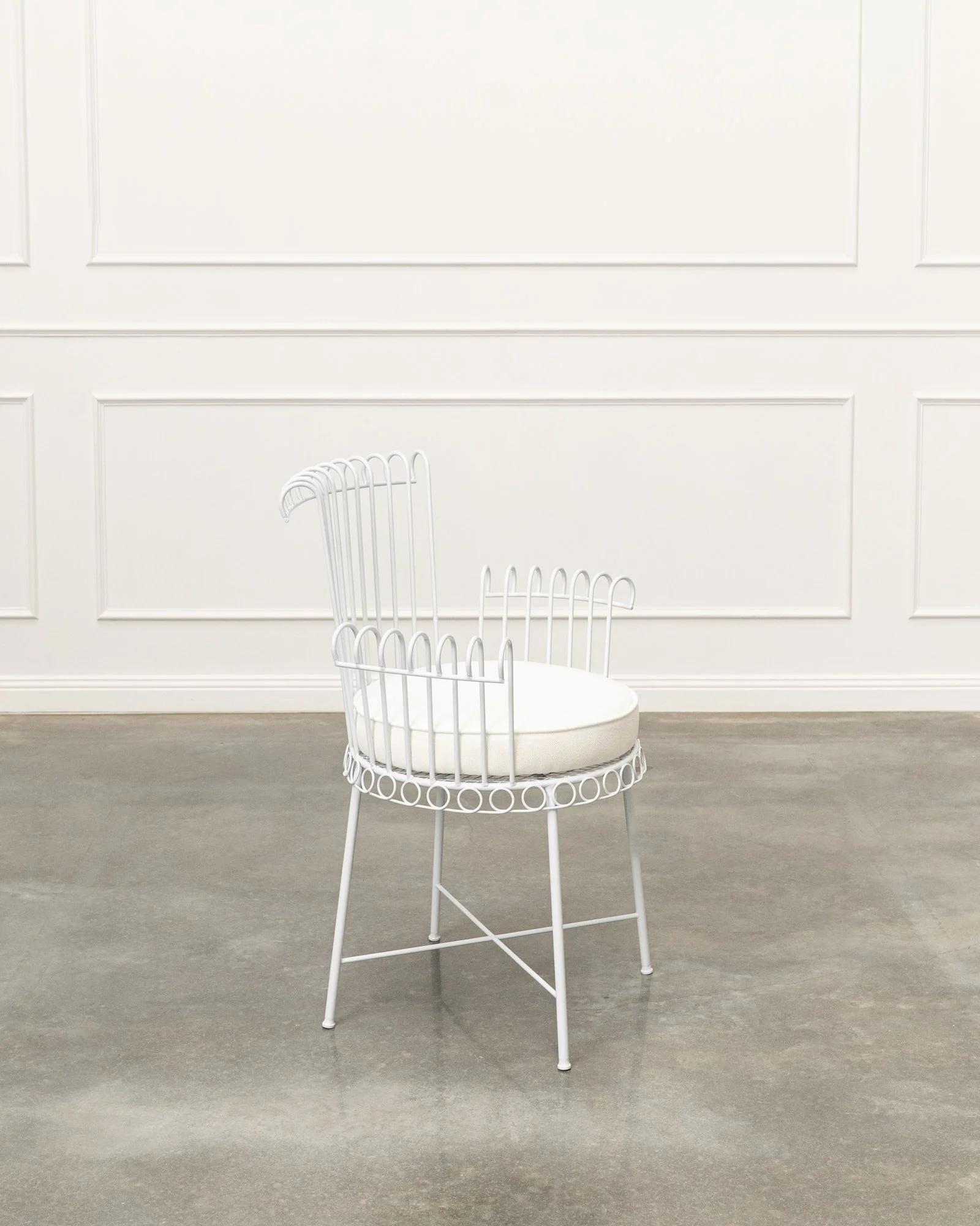 A white wire chair with a circular cushioned seat in a room with white paneled walls and gray flooring.