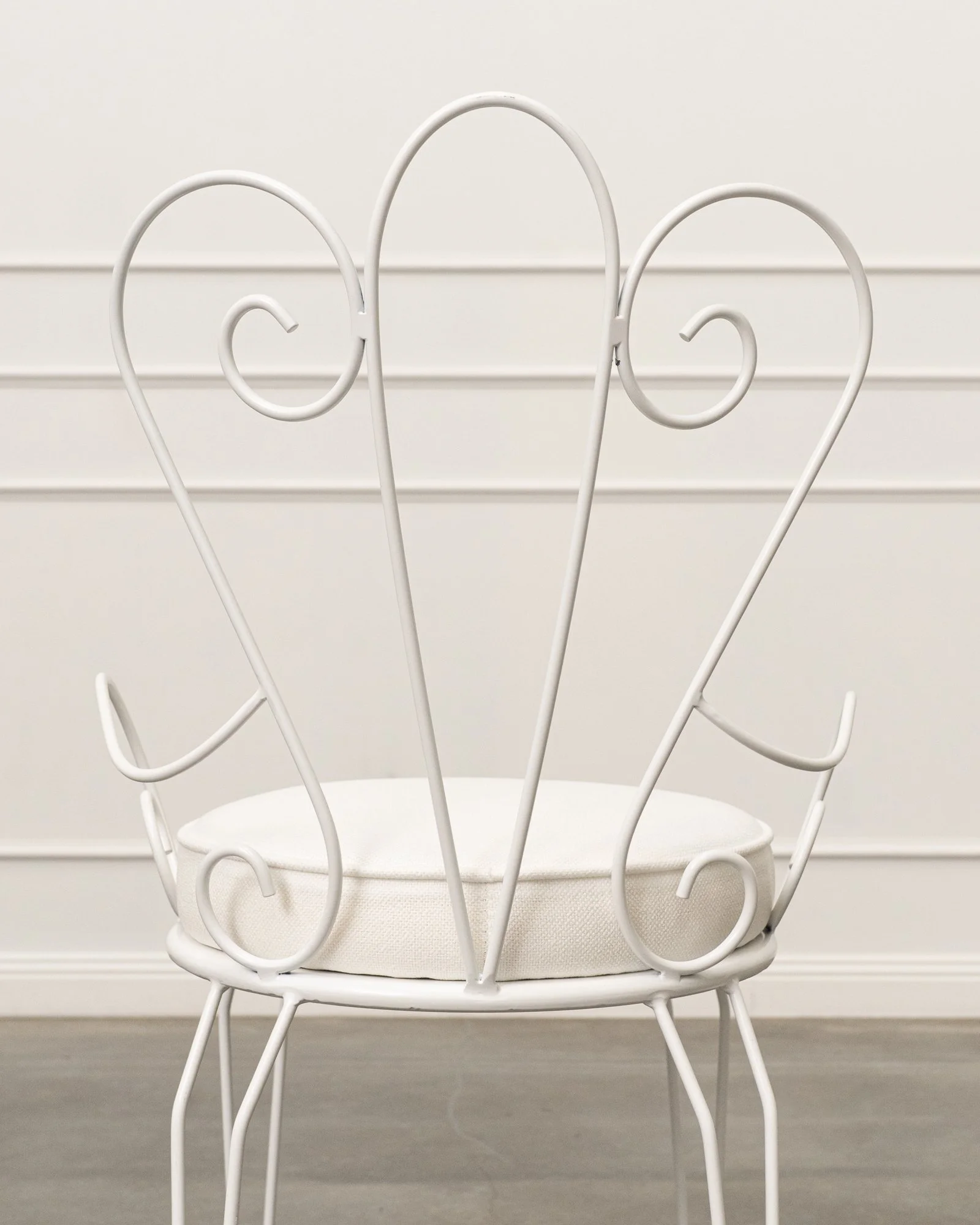 White decorative chair with scrollwork design, positioned in front of a light-colored wall with horizontal molding.