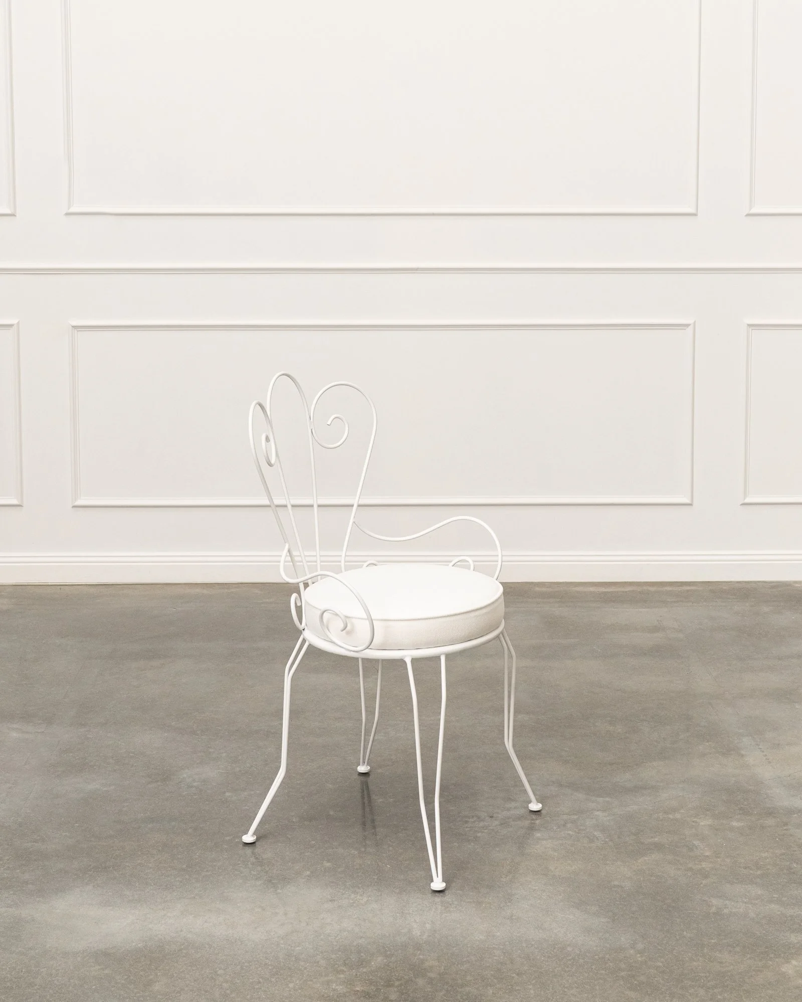 White metal chair with curly decorative elements, placed in front of a white paneled wall.