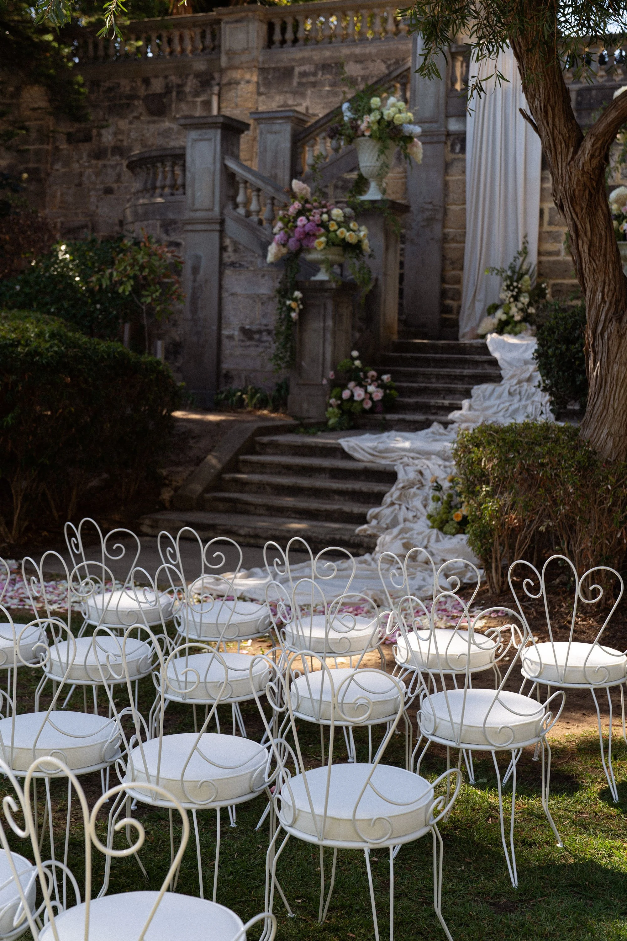 Set of white metal garden chairs arranged around a small round table outdoors, with a backdrop of stone stairs decorated with flowers, draped white fabric, and greenery, suggesting a wedding or special event setting.