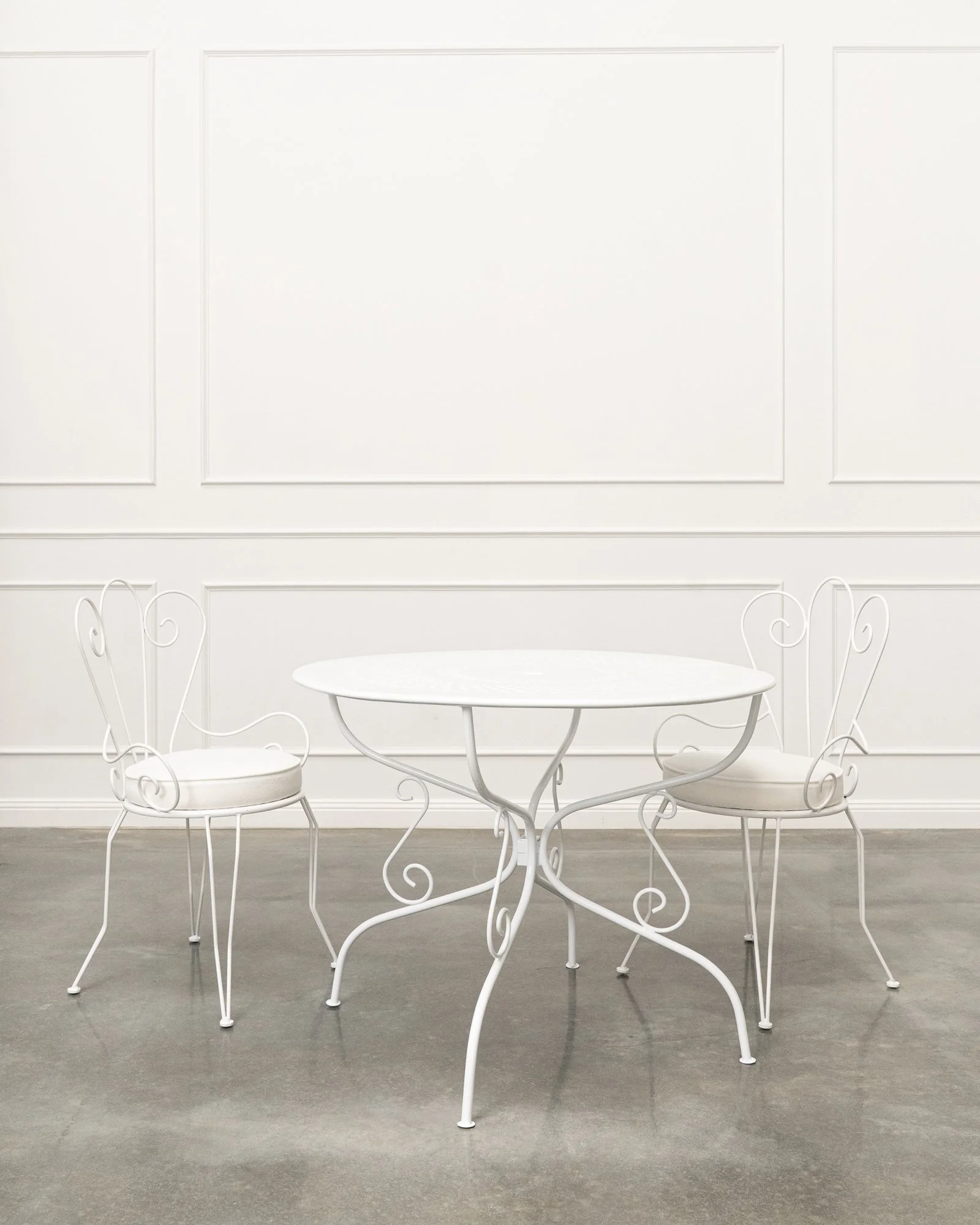 White metal table and two matching chairs with ornate curls in a room with white paneled walls and gray floor.