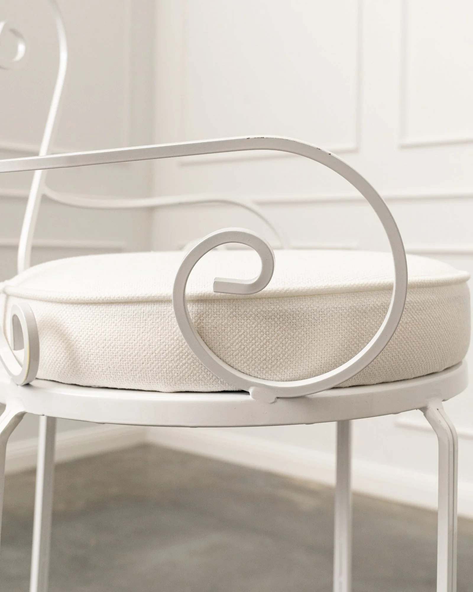 A close-up of a white metal bed frame with curved details, placed on a white cushion or mattress, against a white paneled wall.