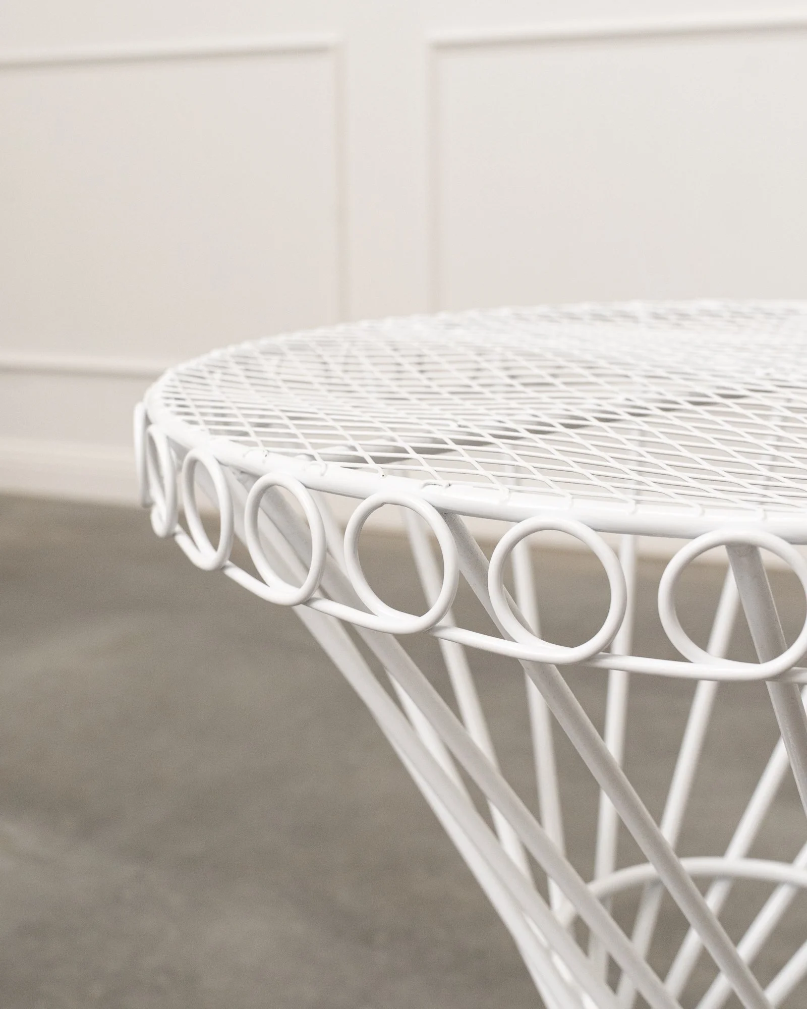Close-up of white wire mesh table with circular legs in front of beige wall with subtle paneling.