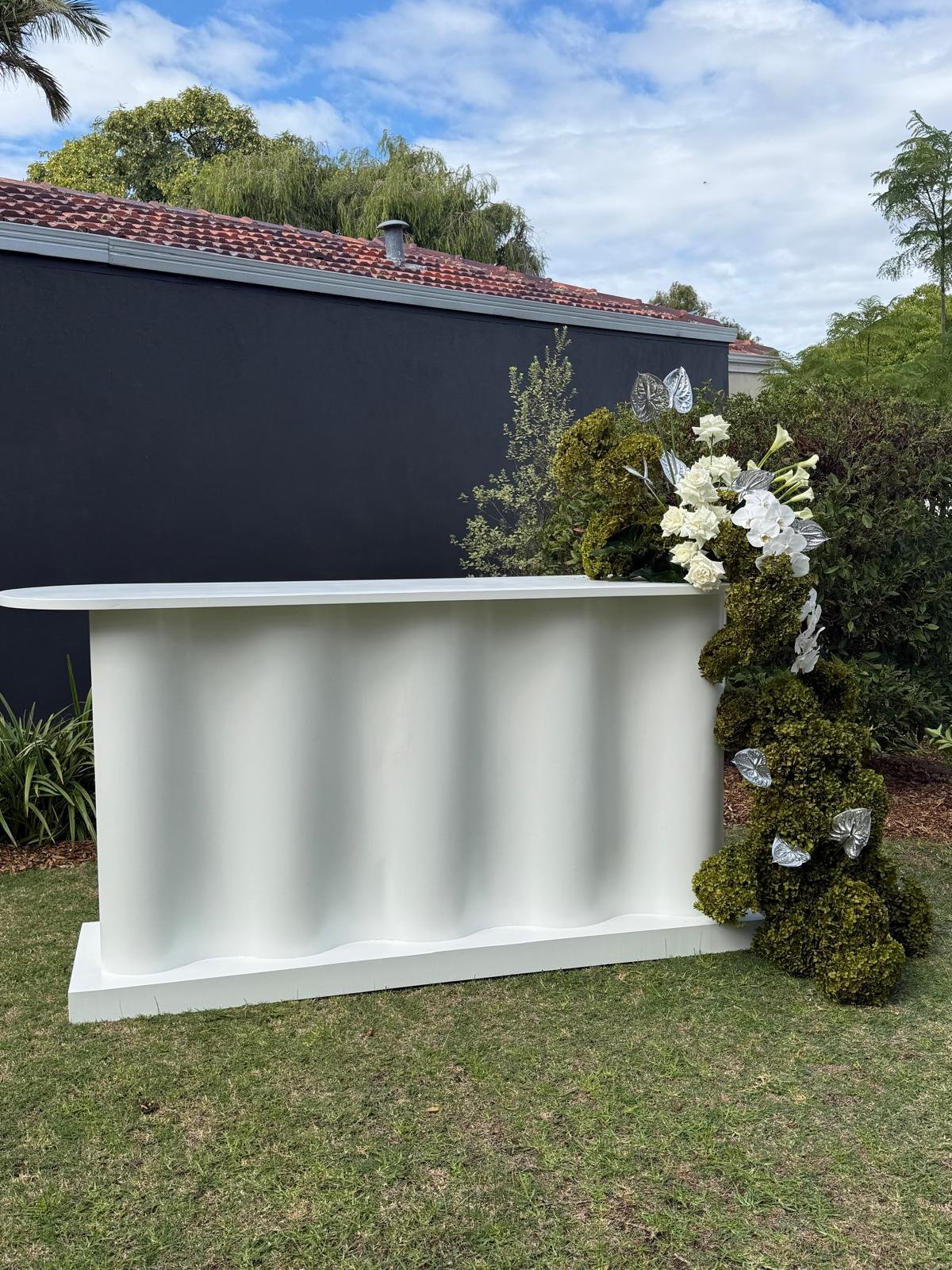 White bar or counter with curved edges, outdoors on grass, decorated with white and green floral arrangements on the right side, set against a dark gray wall, with trees and a partly cloudy sky in the background.