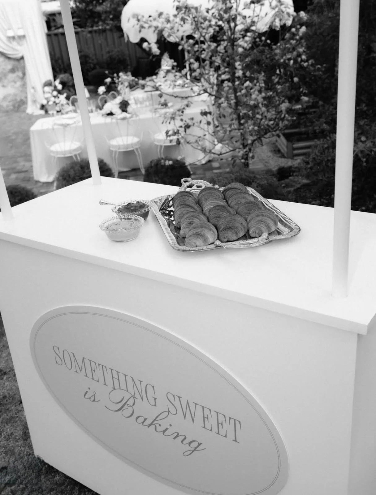 A white bakery cart with a sign that reads "Something Sweet is Baking." On top of the cart, there is a silver tray with croissants and a small bowl of jam or butter.