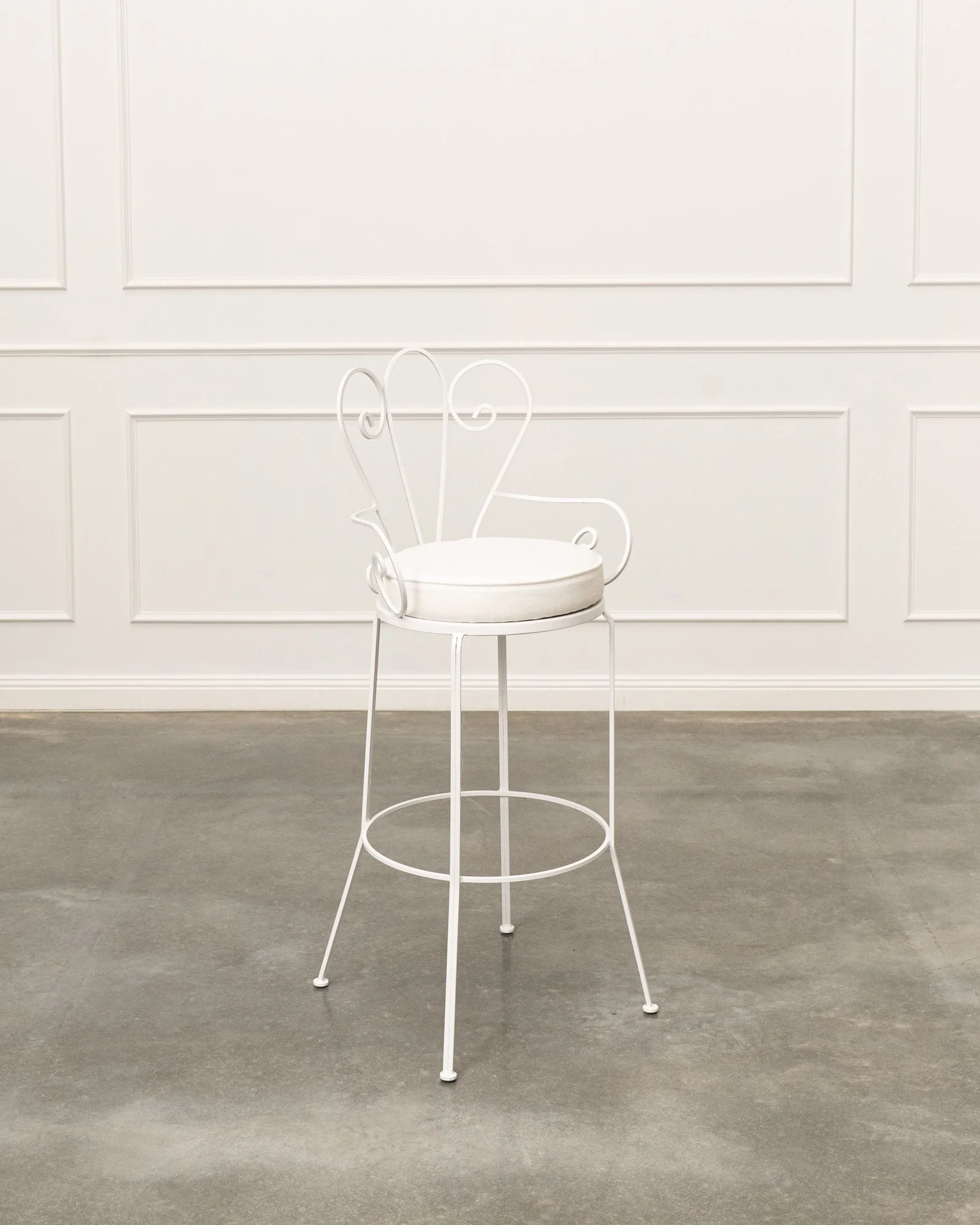 White metal chair with heart-shaped backrest and round cushion seat, in front of a white paneled wall with gray floor.
